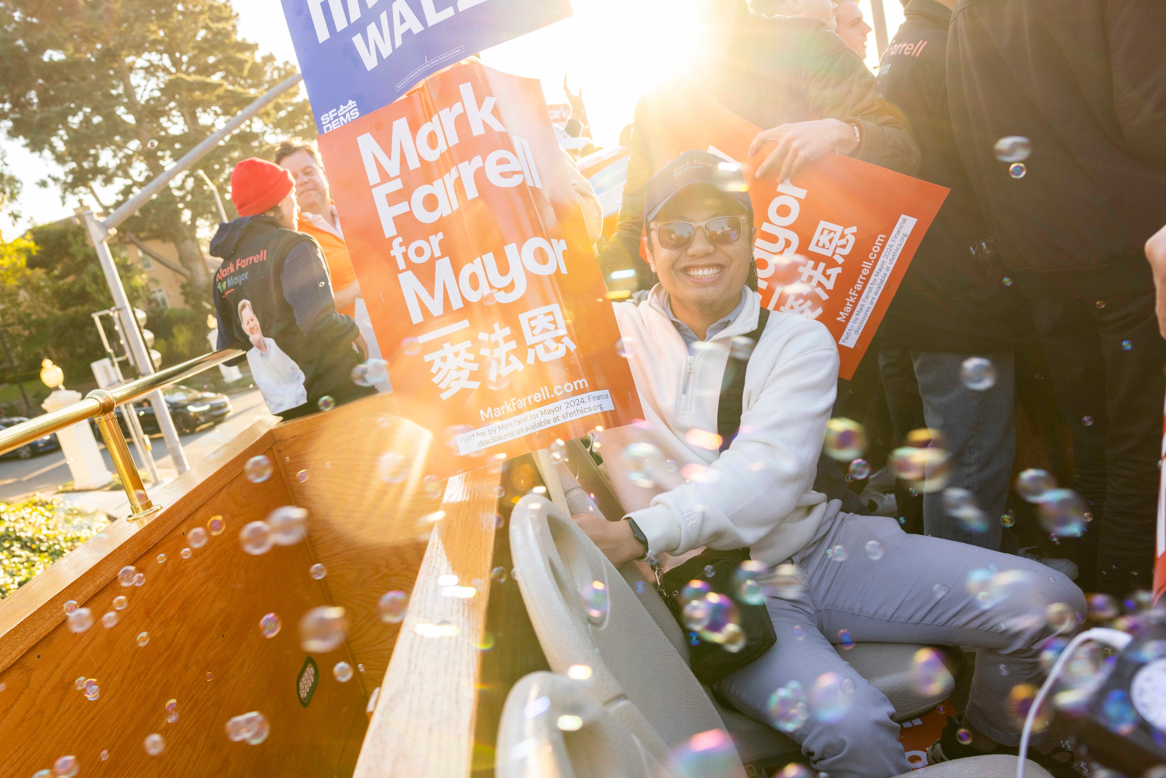 A smiling person in sunglasses holds a &quot;Mark Farrell for Mayor&quot; sign while sitting on a vehicle surrounded by sunlight and soap bubbles.