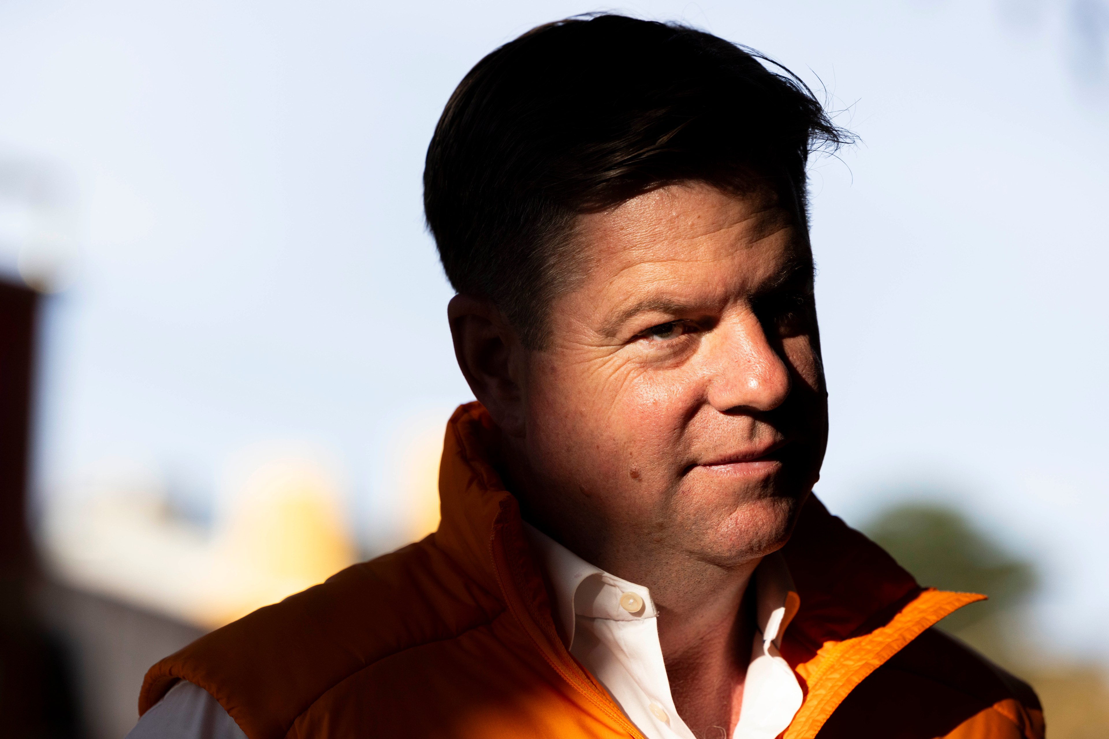 A man with short dark hair wears an orange jacket over a white shirt, standing against a blurred background. His face is partially lit, with a subtle smile.