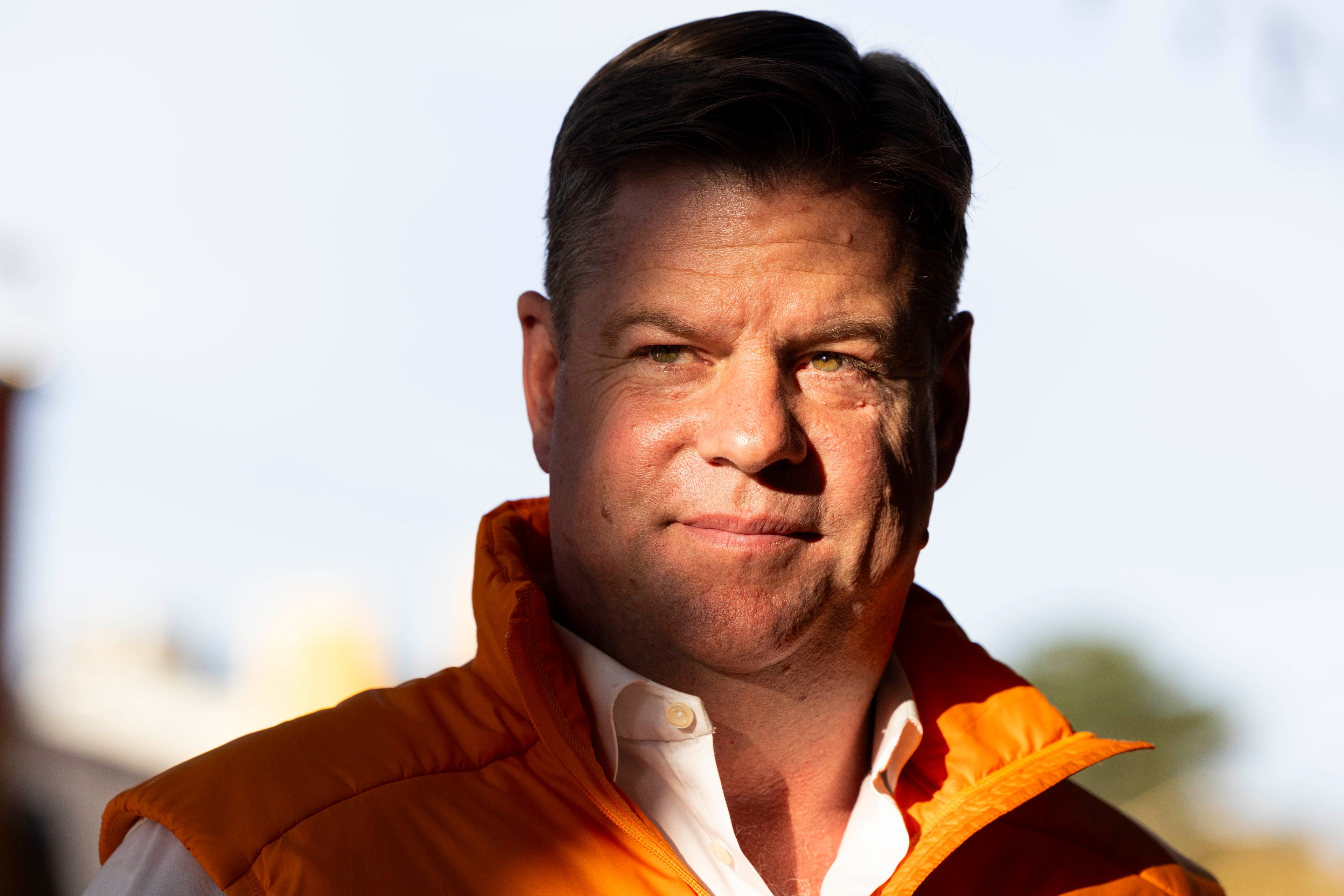 A man is shown in partial sunlight, wearing an orange jacket over a white shirt. His expression is neutral, and the background is blurry.