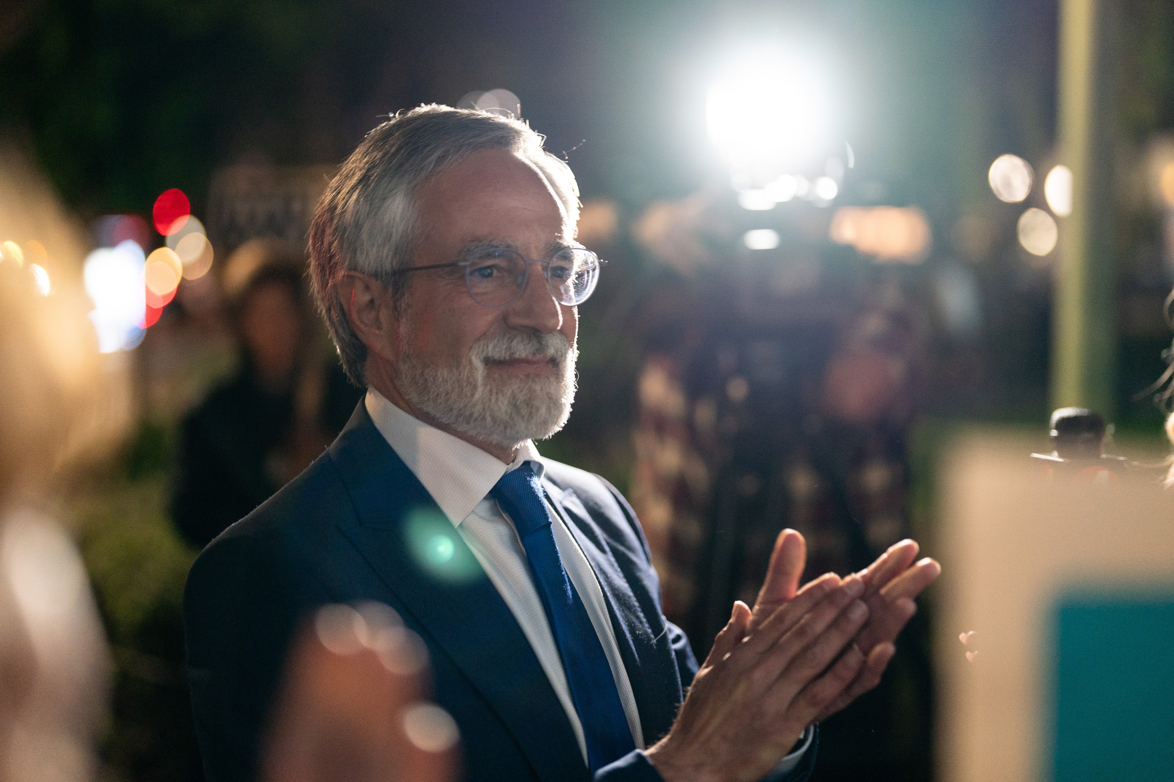 A man with glasses and a beard is smiling in a suit, clapping under bright lights, with a blurred crowd and camera in the background.