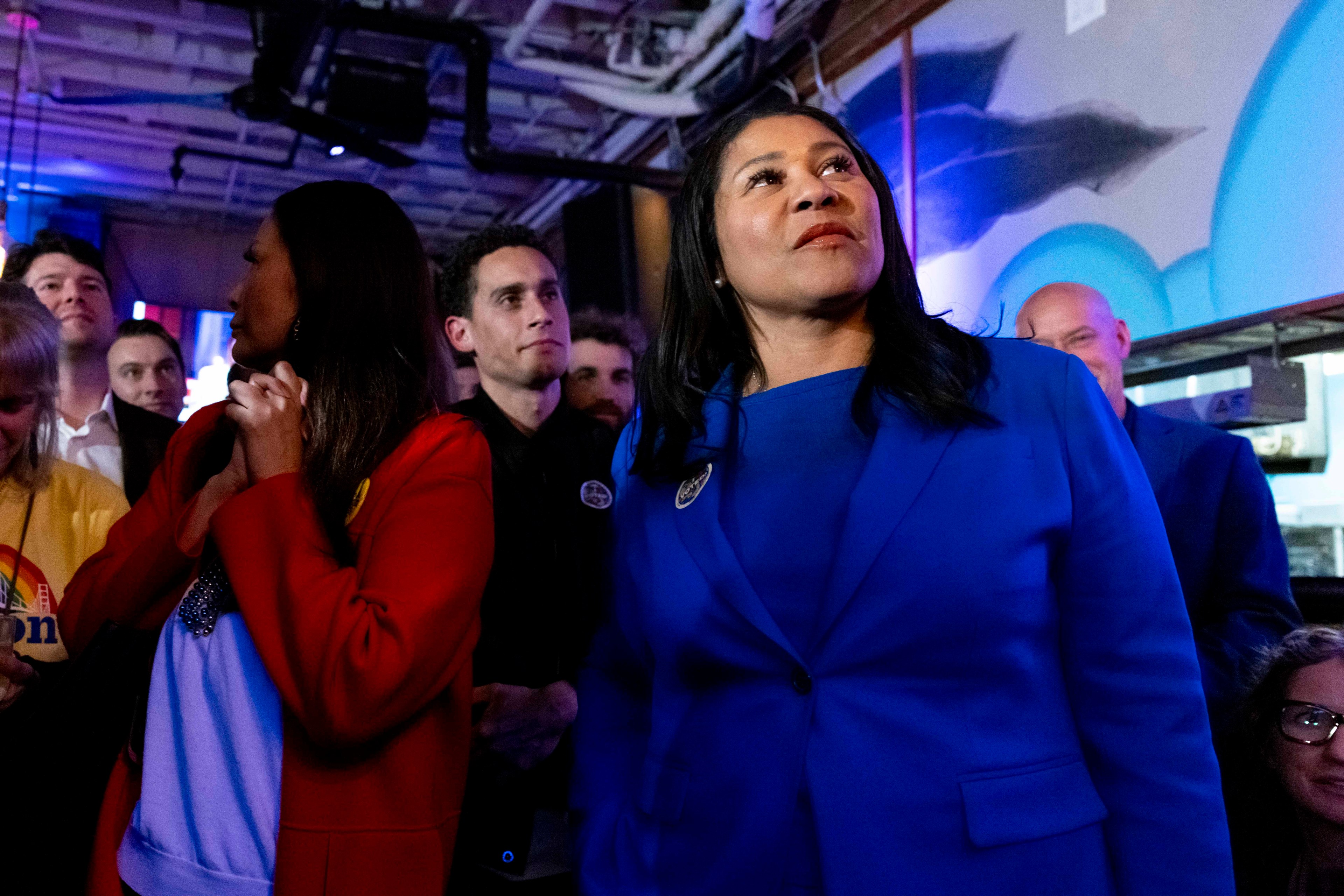 A woman in a blue blazer stands confidently under colorful lighting, surrounded by a group of people, some in casual attire, in a dimly lit room.