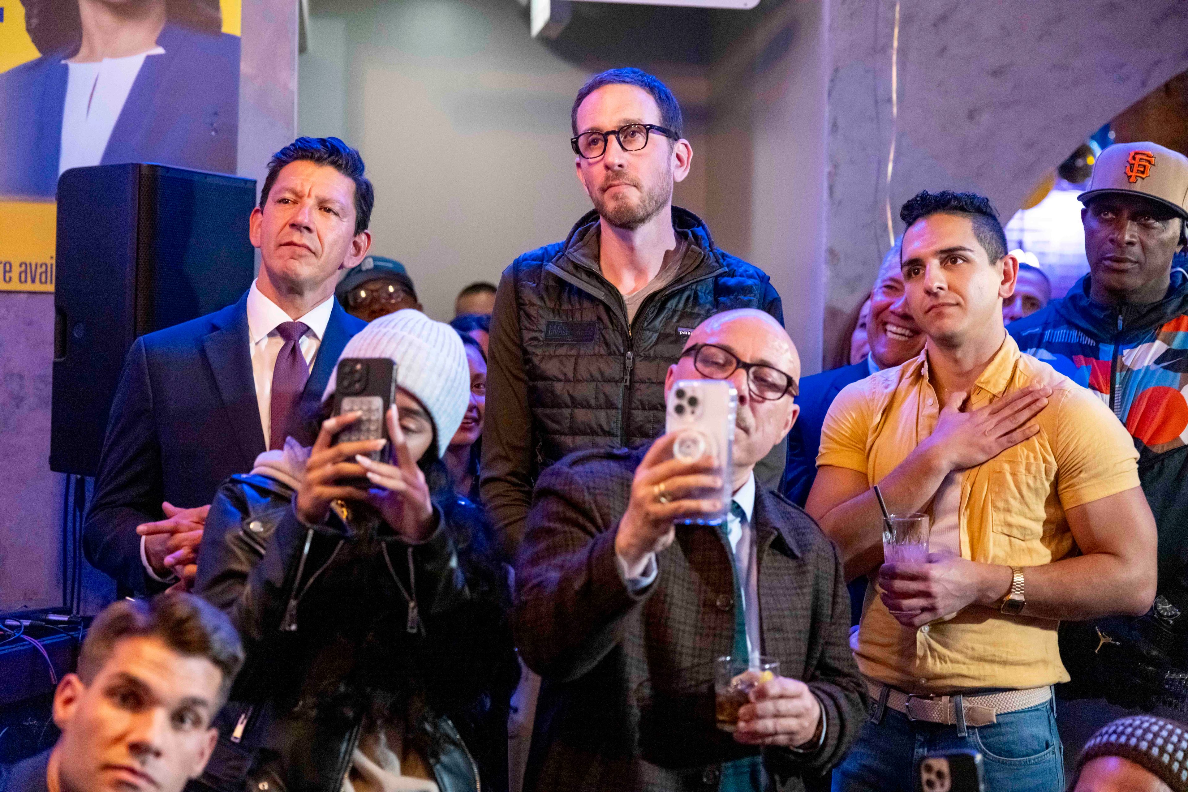 A group of people are gathered indoors, some taking photos with phones. They appear attentive and engaged, with a mix of casual and formal attire.