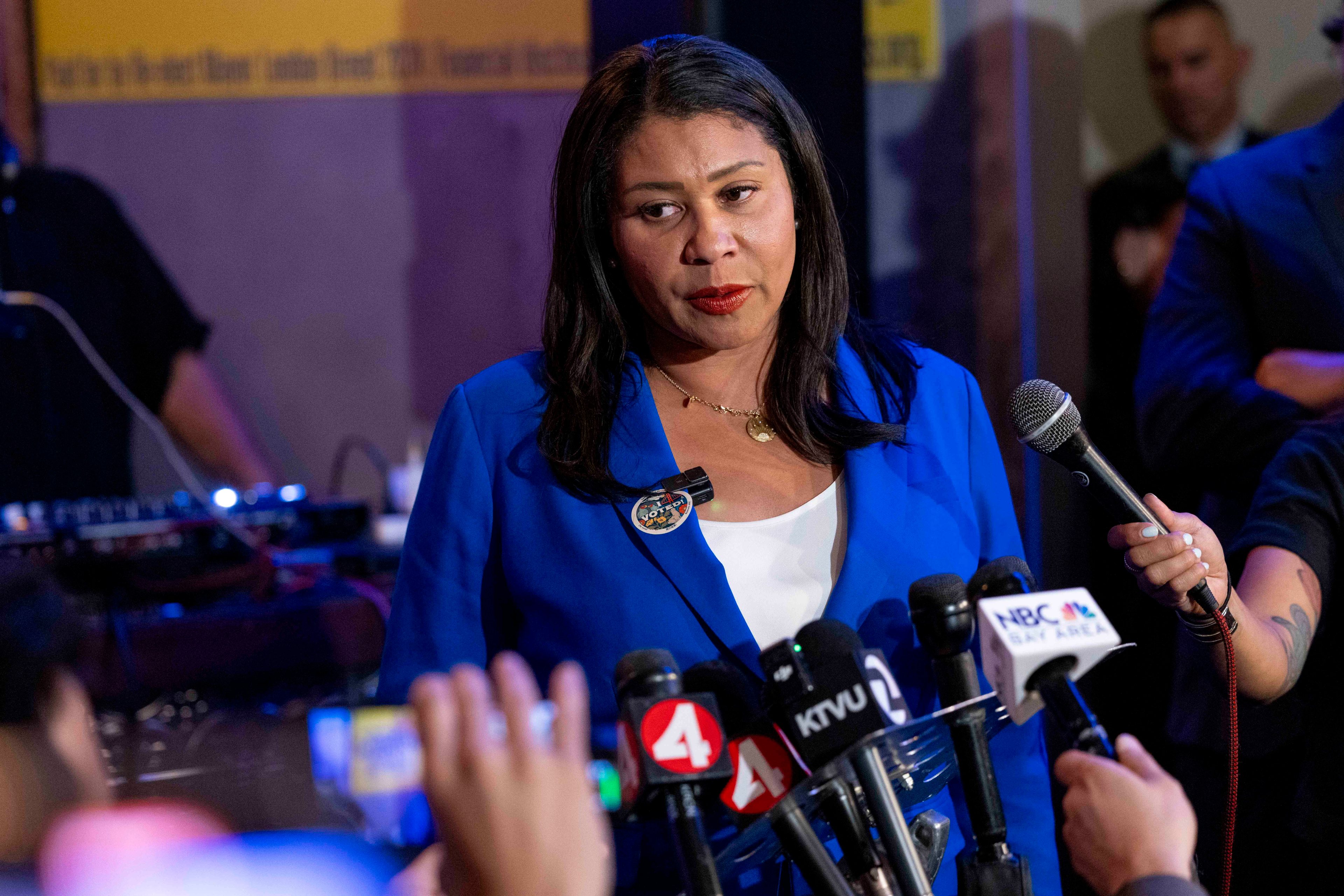 A woman in a blue blazer stands before a cluster of microphones from various news outlets, addressing the media with a serious expression.