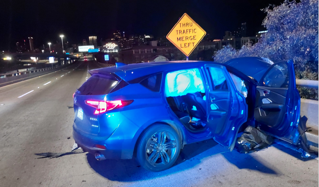 A blue car with deployed airbags and open doors is on a highway at night. Its front is damaged, and a "Thru Traffic Merge Left" sign is visible nearby.