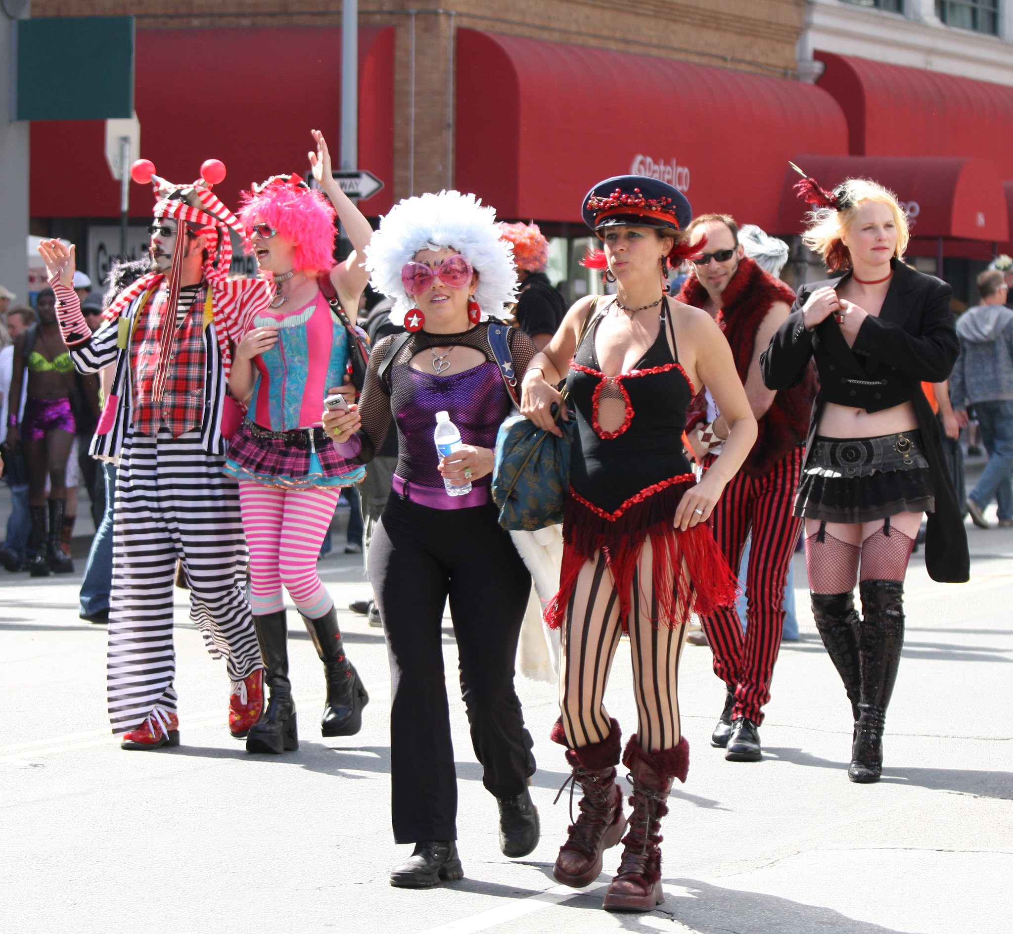 A group of people is walking in a parade, dressed in colorful, eccentric costumes with vibrant wigs, striped patterns, and theatrical accessories.