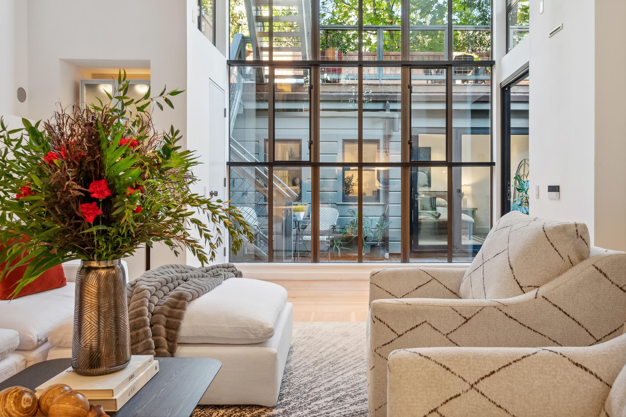 A cozy living room with plush chairs, a side table with a vase of red flowers, and a knitted throw blanket. Large windows offer a view of the outside greenery.