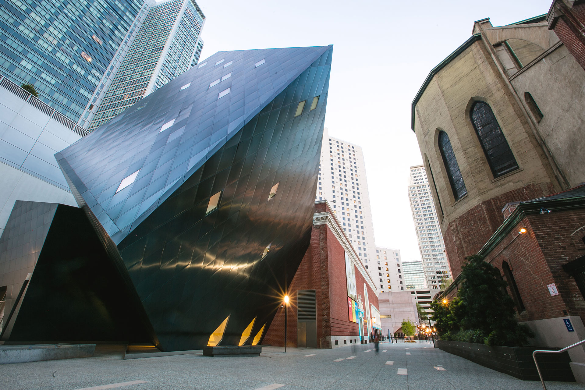 The image shows a modern, angular black building next to a traditional brick structure. Tall skyscrapers rise in the background on a clear day.