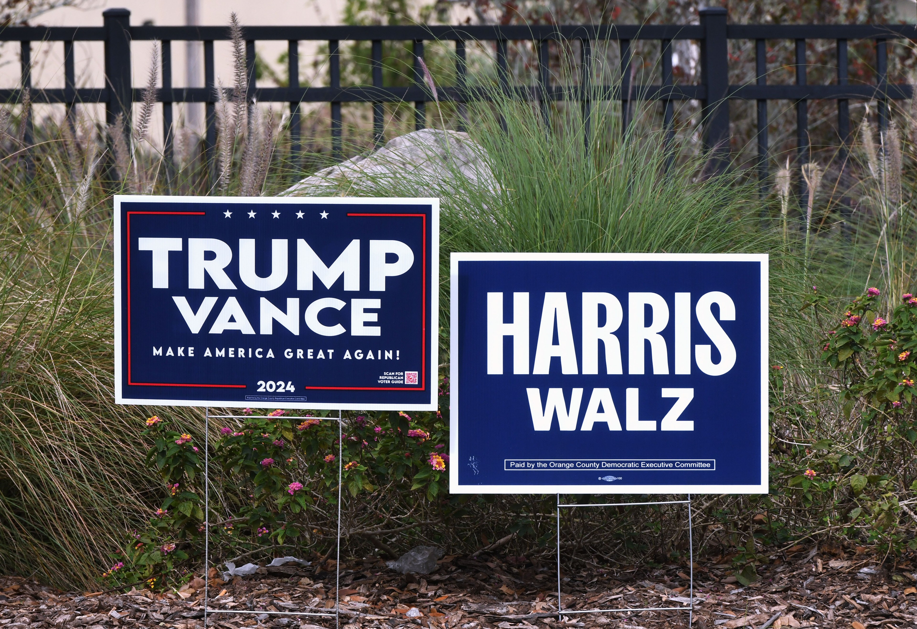 Two political yard signs are in front of grass and flowers: one for &quot;Trump Vance 2024&quot; with &quot;Make America Great Again!&quot; and another for &quot;Harris Walz.&quot;