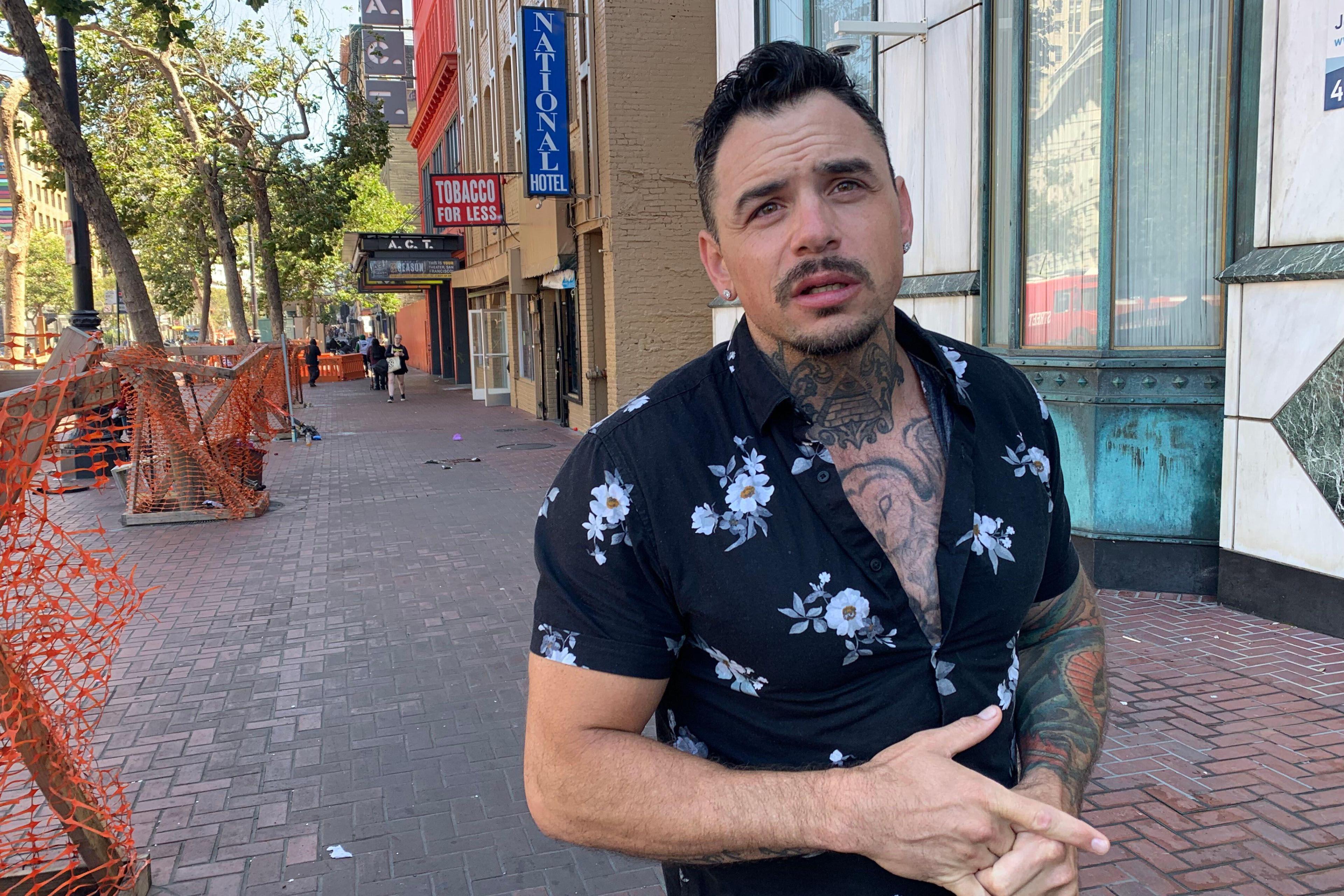 A man with tattoos, wearing a floral shirt, stands on a brick sidewalk. Behind him are trees, orange fencing, and storefronts, including a "Tobacco for Less" sign.