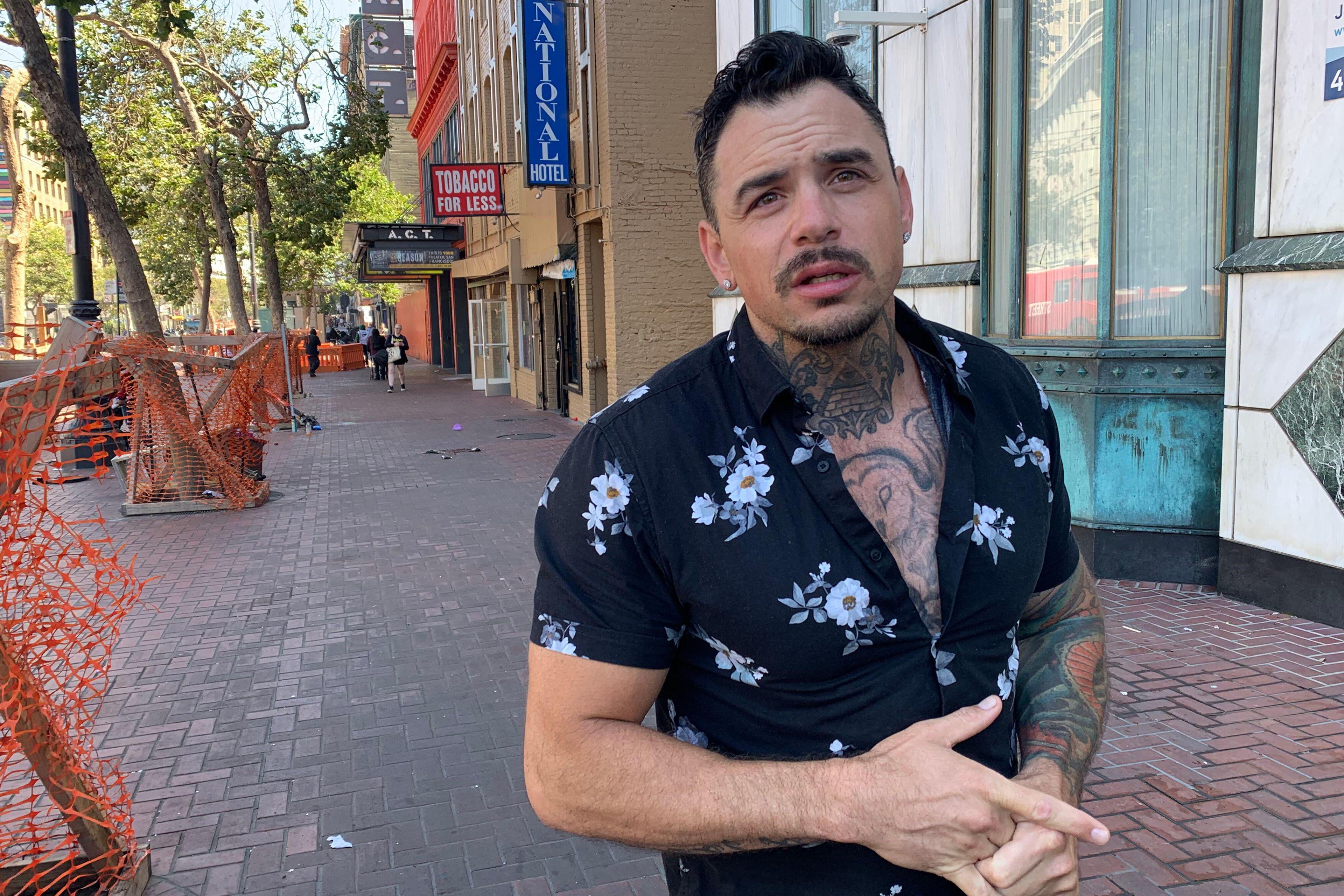 A man with tattoos, wearing a floral shirt, stands on a brick sidewalk. Behind him are trees, orange fencing, and storefronts, including a "Tobacco for Less" sign.