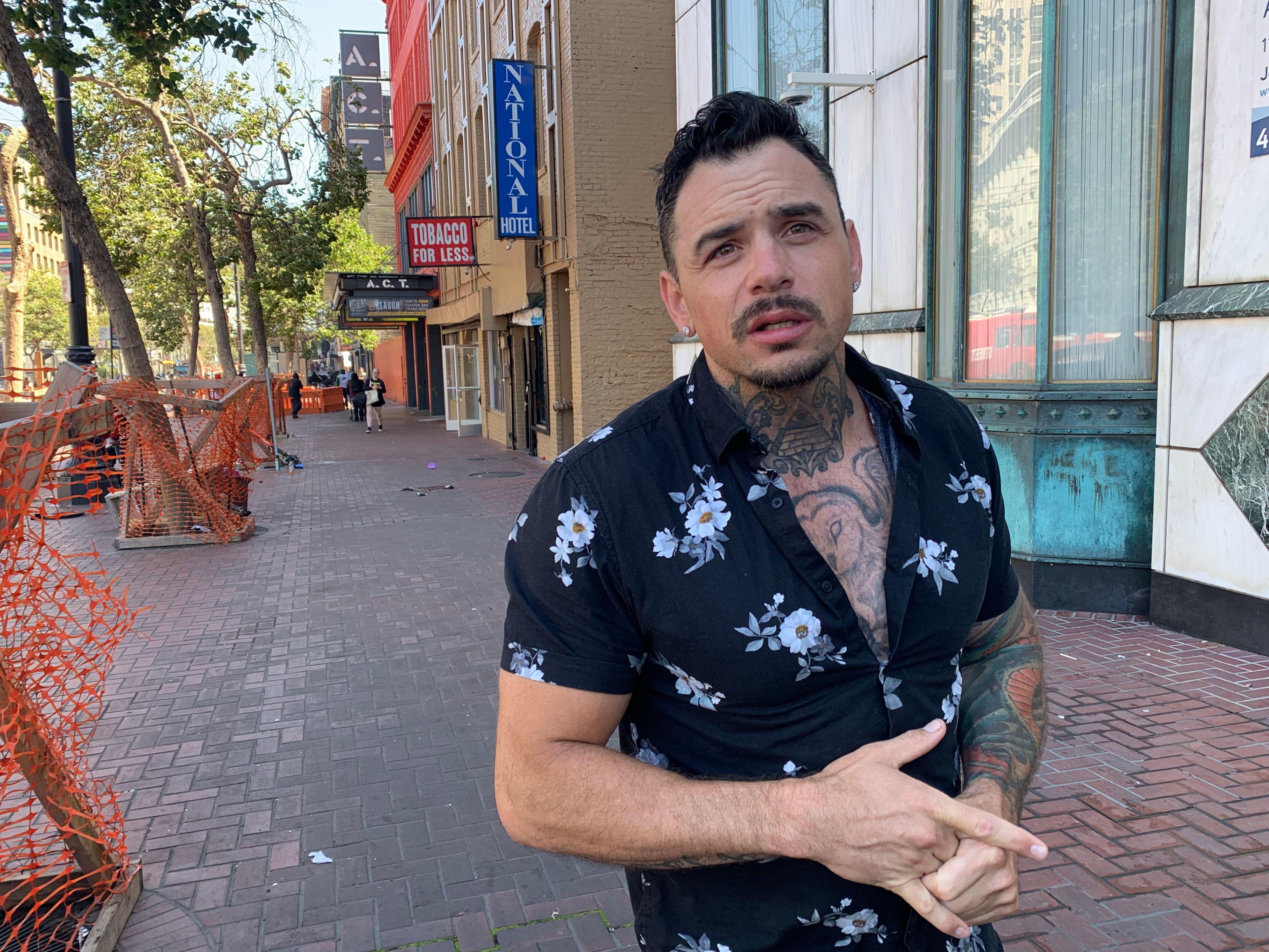 A man with tattoos, wearing a floral shirt, stands on a brick sidewalk. Behind him are trees, orange fencing, and storefronts, including a "Tobacco for Less" sign.