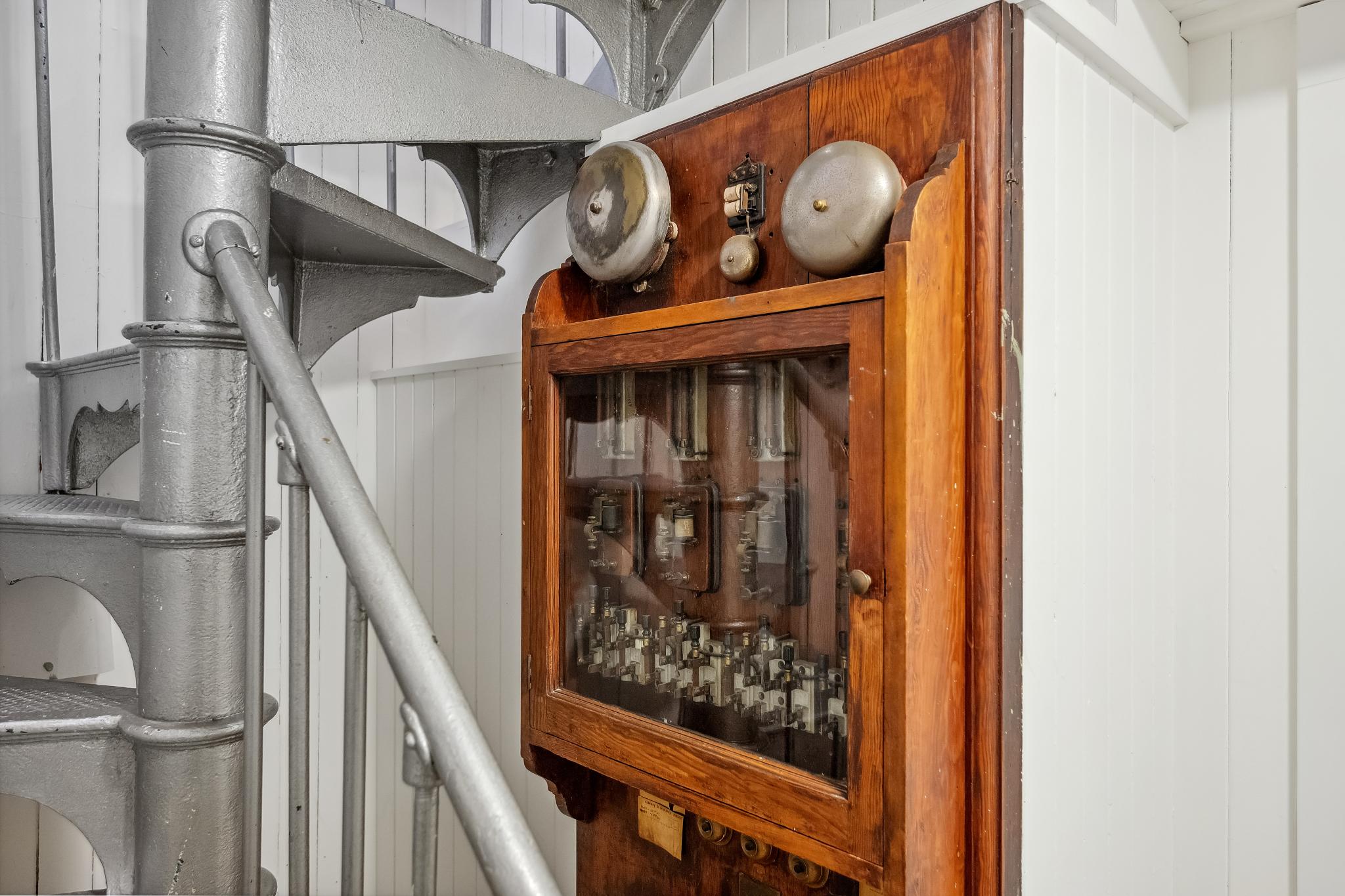 A metal spiral staircase is beside a wooden, glass-fronted electrical panel with gauges and knobs. The wall behind is white with vertical lines.