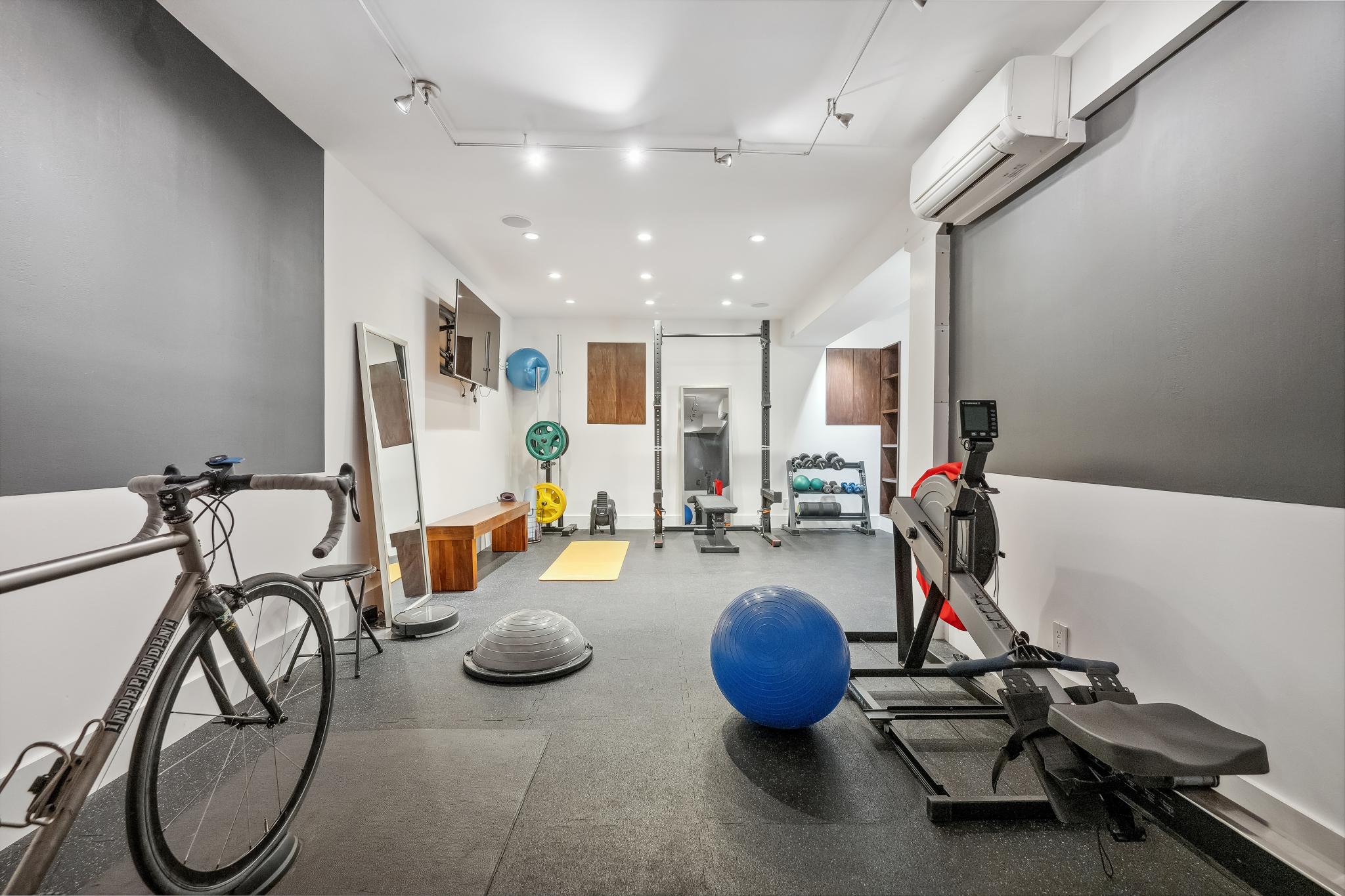 A home gym with a bike, rowing machine, blue stability ball, bench, free weights, and wall mirror. There's also a wall-mounted TV and various exercise equipment.