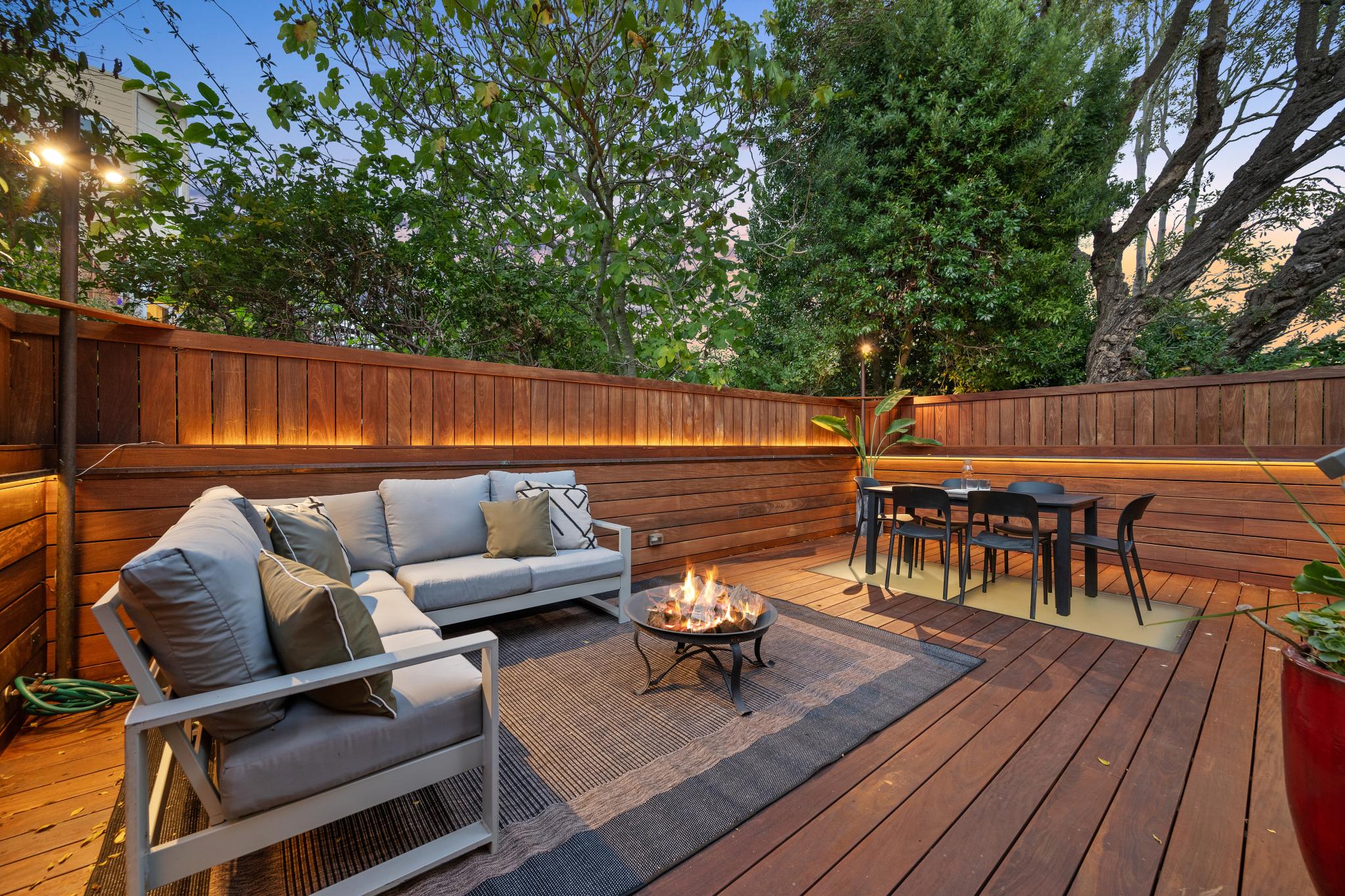 A cozy outdoor patio features a gray sectional sofa, a fire pit, and a dining table. Wooden fencing with greenery surrounds the space, illuminated by soft lights.