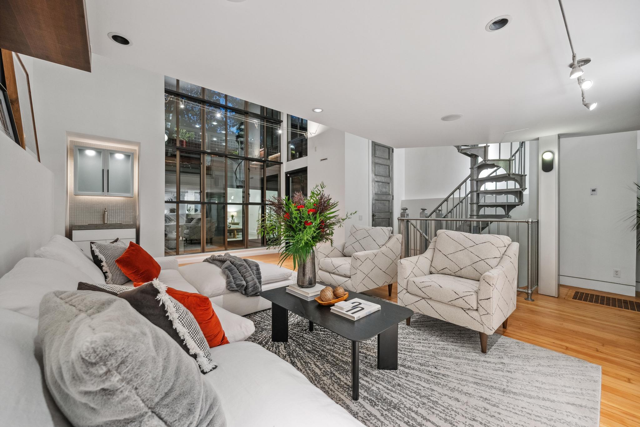 A modern living room with a white sofa, two patterned armchairs, a coffee table with flowers, and a spiral staircase, all set against large glass windows.