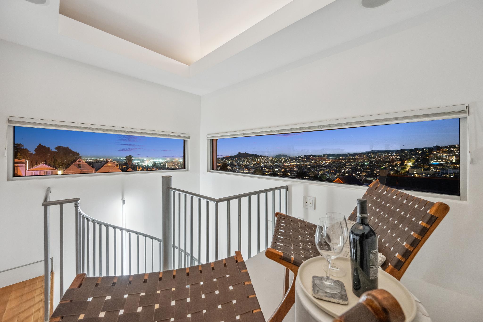 Two chairs and a round table with wine and glasses overlook a cityscape through wide windows at night, with a spiral staircase on the side.