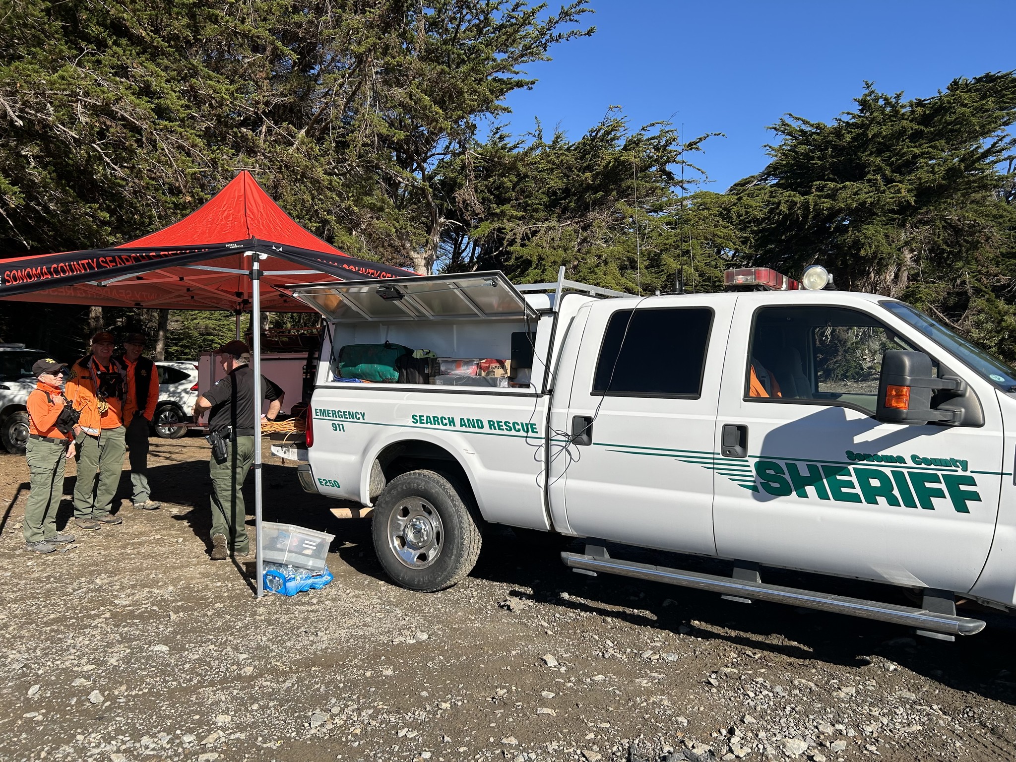 A white sheriff's truck is parked at a search and rescue scene with a red tent and four uniformed personnel gathered nearby under trees.