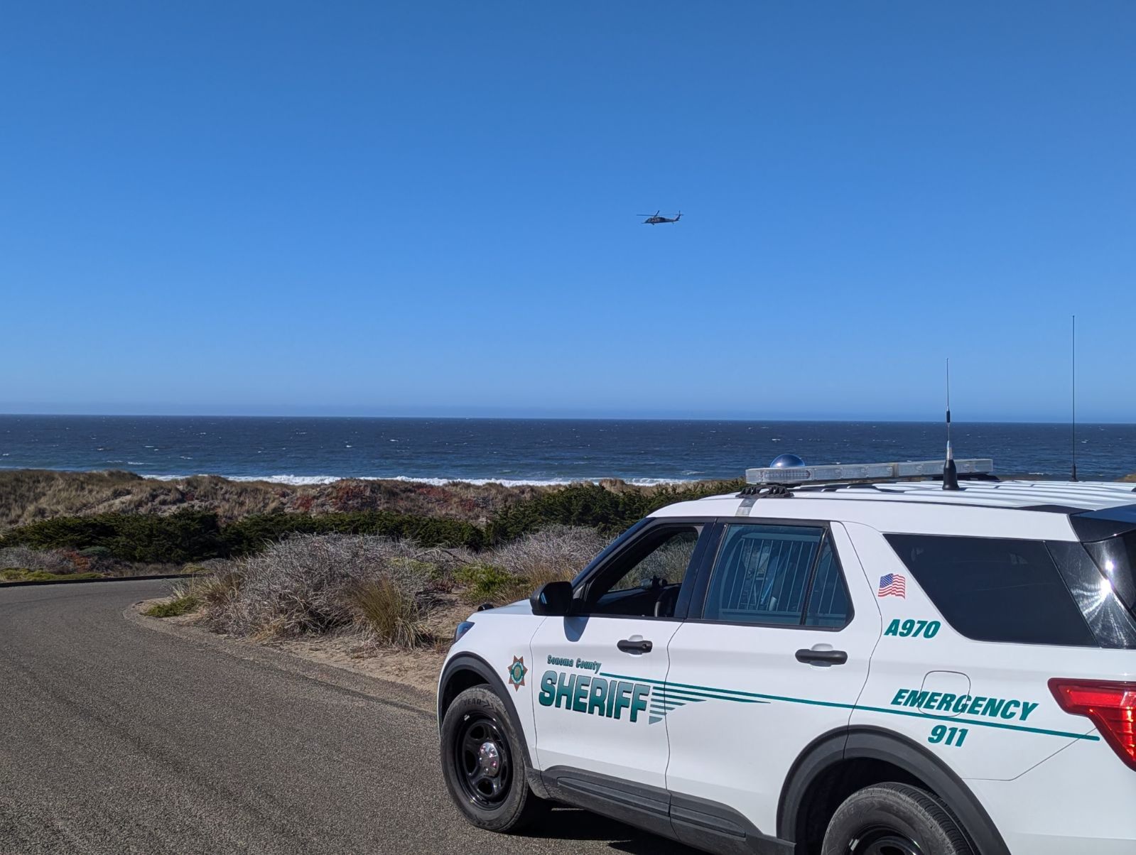 A sheriff's SUV is parked on a coastal road with the ocean and a helicopter in the blue sky above.