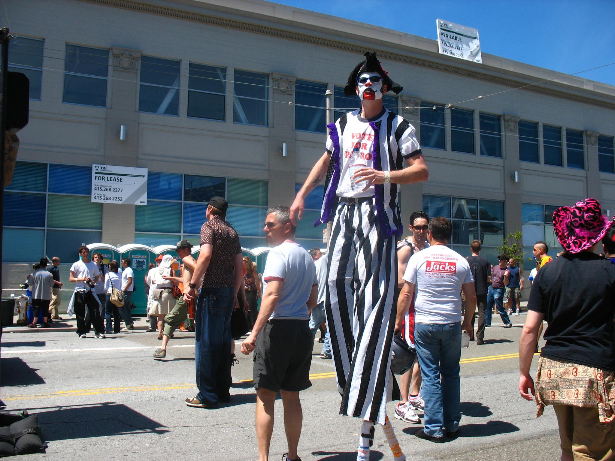 A person on stilts wearing a black-and-white striped outfit and clown makeup stands among a crowd on a sunny street, with a building in the background.