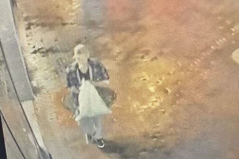 A person walks on a wet street at night, carrying a white plastic bag. The ground reflects streetlights, and several figures can be seen in the background.