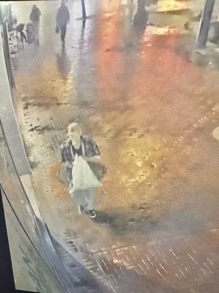 A person walks on a wet street at night, carrying a white plastic bag. The ground reflects streetlights, and several figures can be seen in the background.