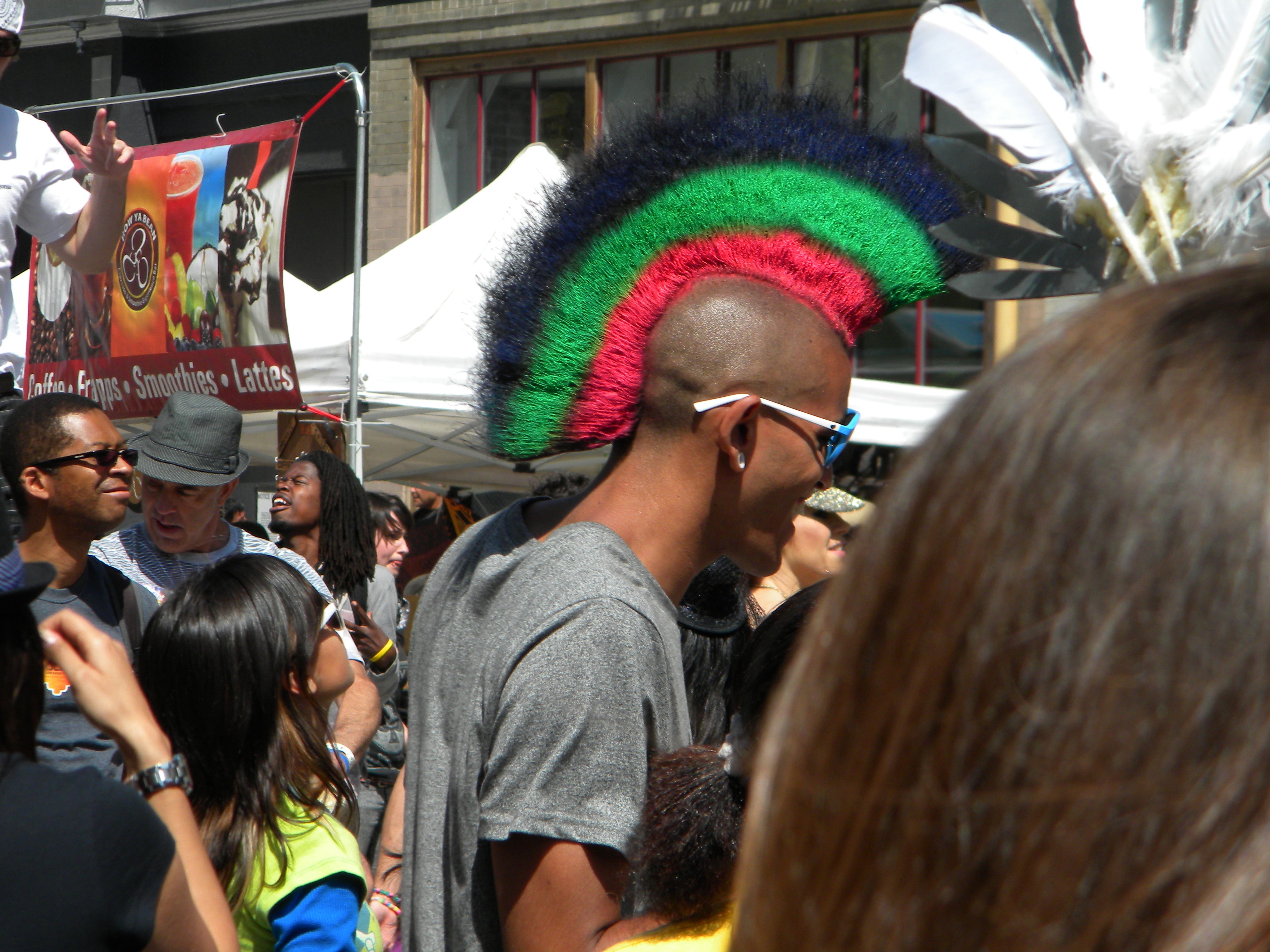 A person with a vibrant multi-colored mohawk and sunglasses is in a crowd during an outdoor event, with various people and a food stand in the background.