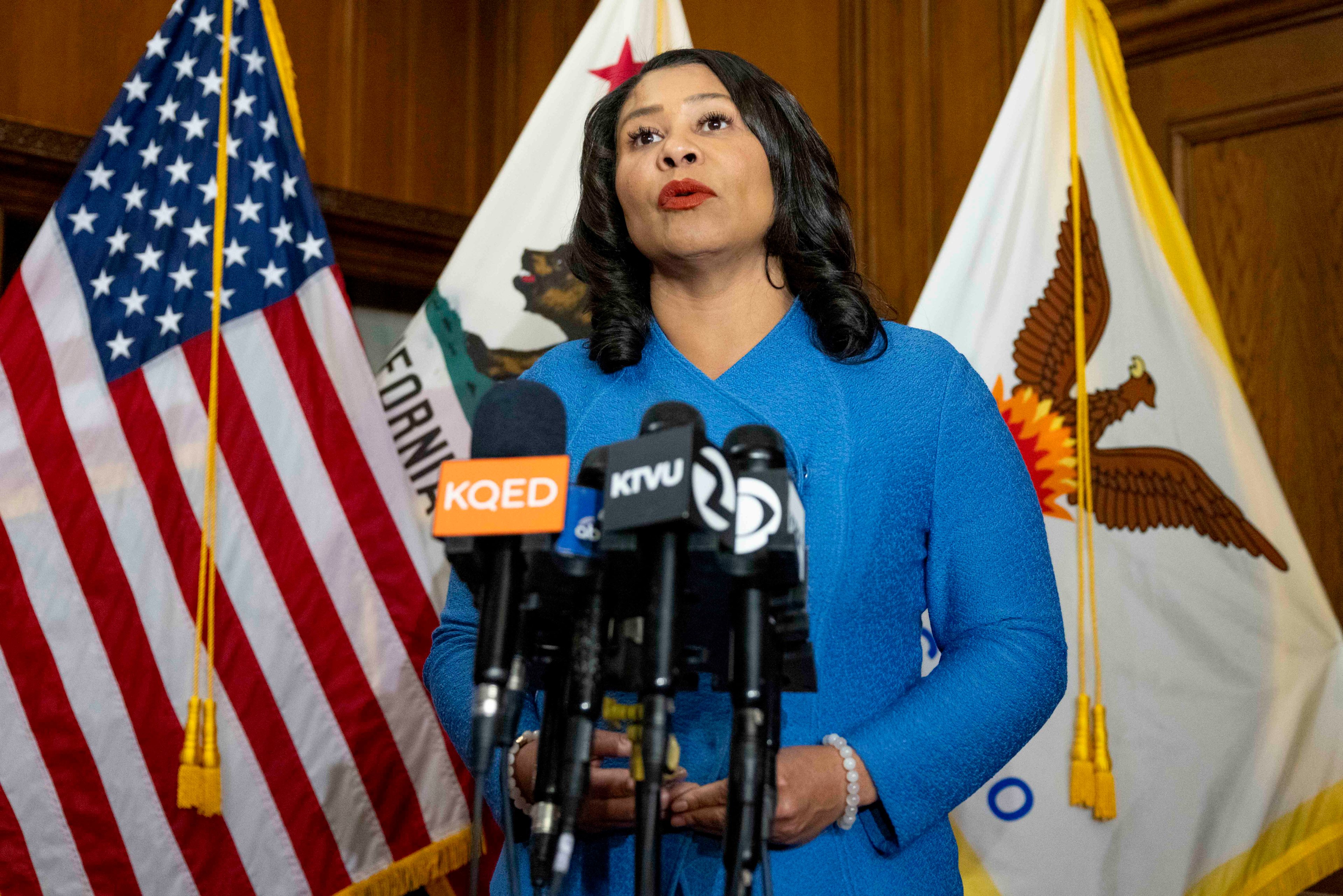 A woman in a blue outfit speaks at a podium with multiple microphones. Behind her are U.S., California, and city flags.