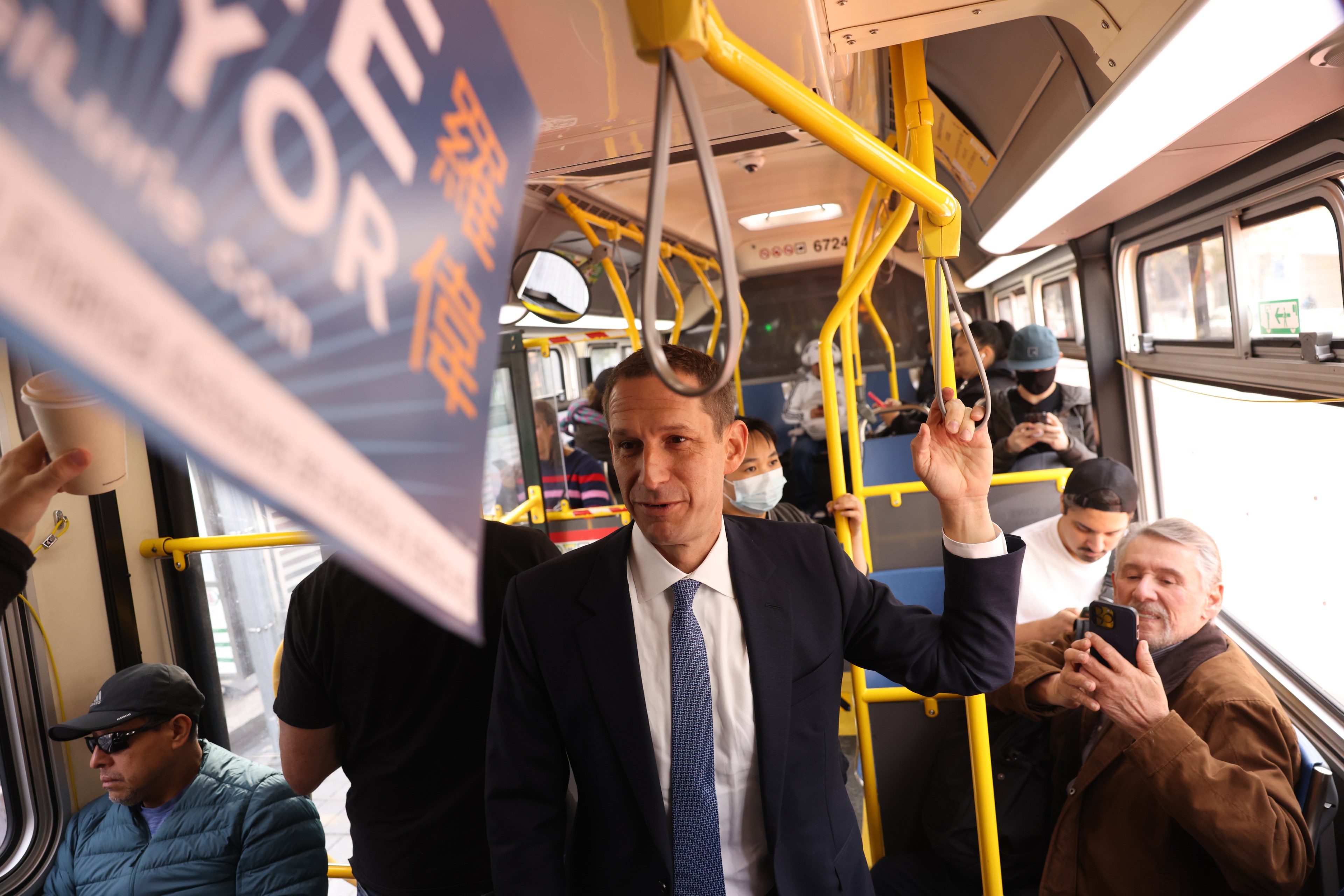 A well-dressed man stands inside a bus, holding onto a rail. Passengers sit and look at their phones, while a campaign sign is slightly visible overhead.