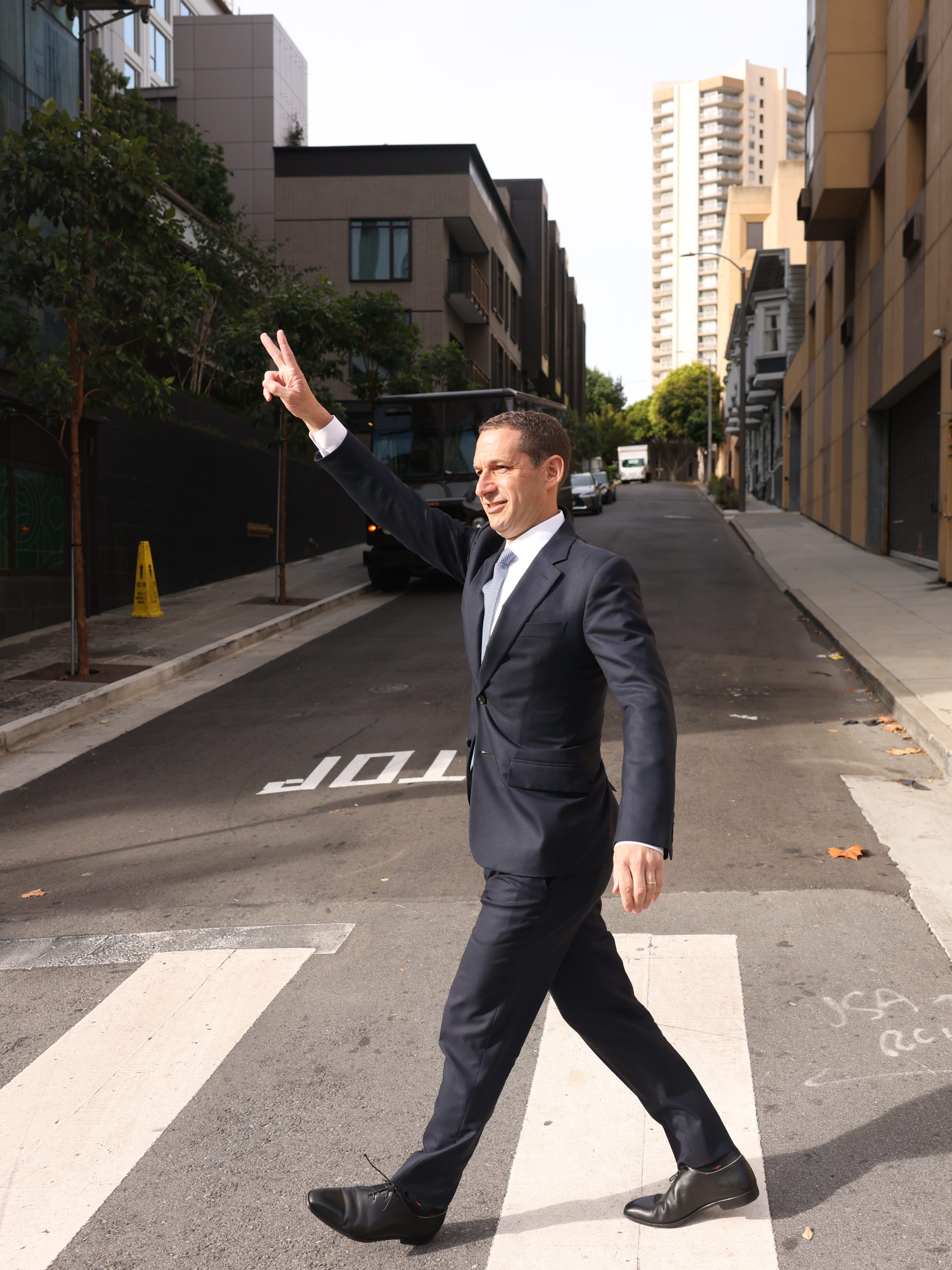 A man in a suit crosses a street on a sunny day, holding up a peace sign with his left hand. Tall buildings and trees line the street.