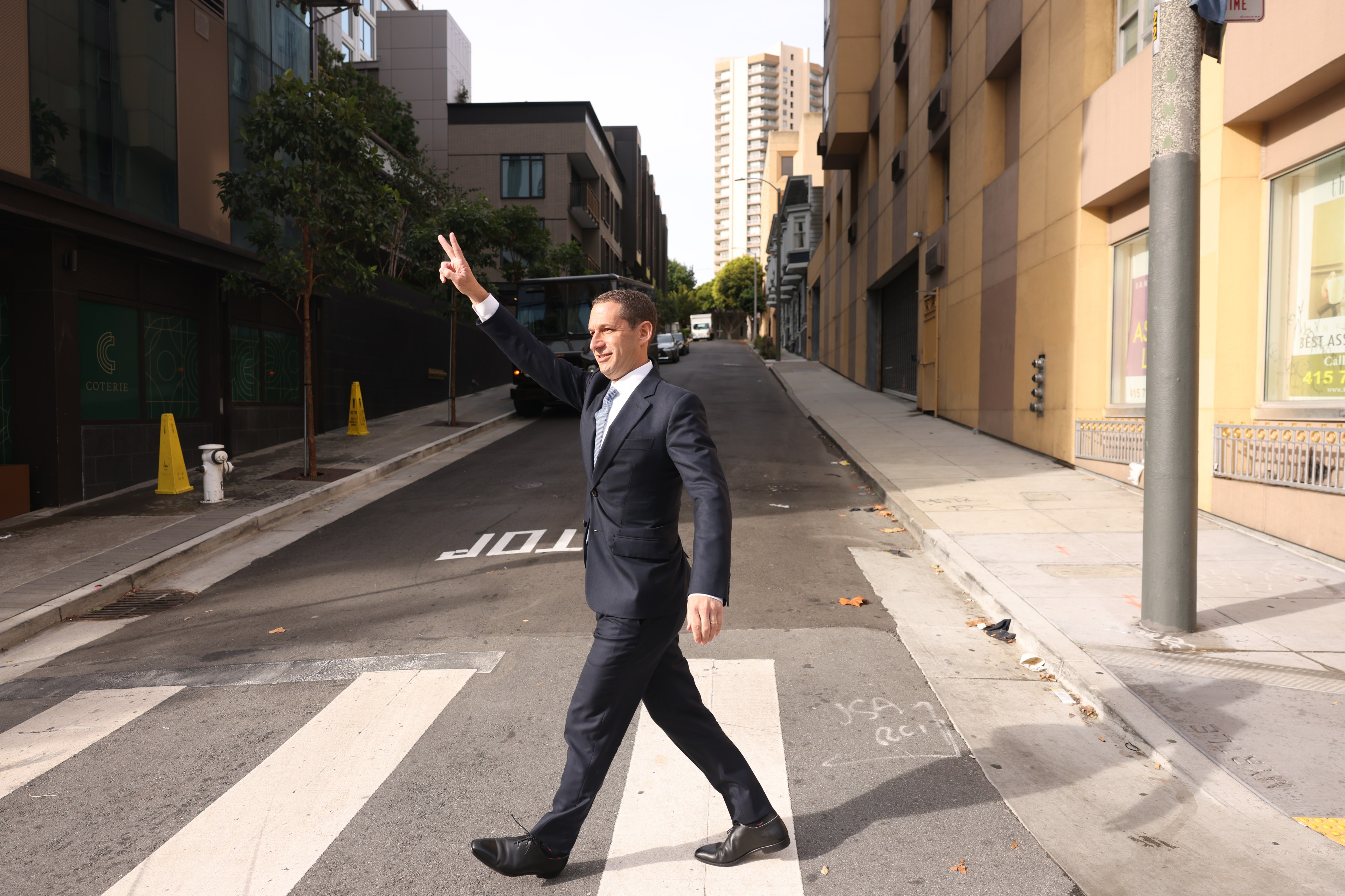 A man in a suit crosses a street on a sunny day, holding up a peace sign with his left hand. Tall buildings and trees line the street.