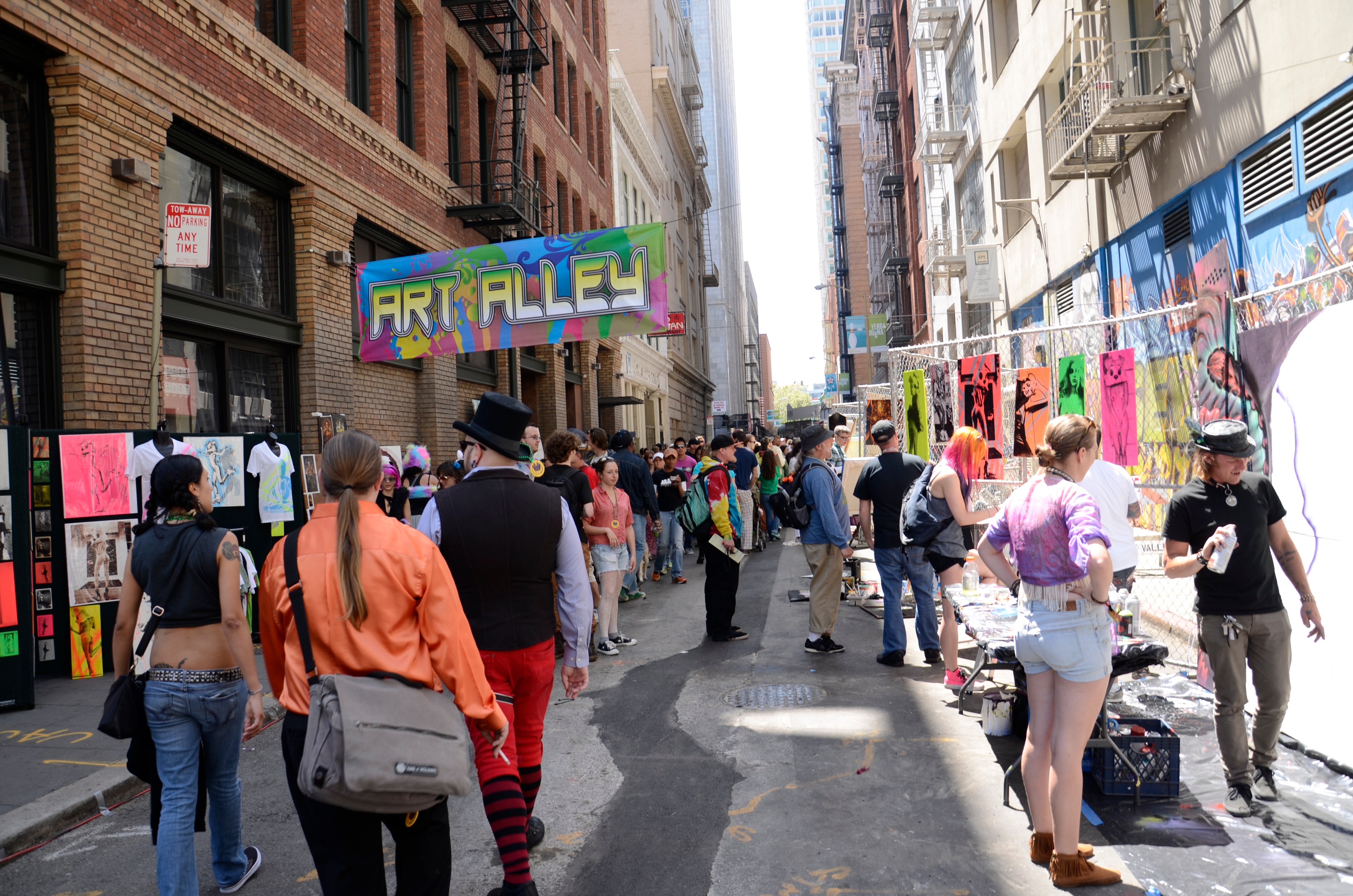 A bustling alley with colorful artwork on display. People walk and interact, some wearing colorful outfits. A vibrant &quot;Art Alley&quot; sign hangs overhead.