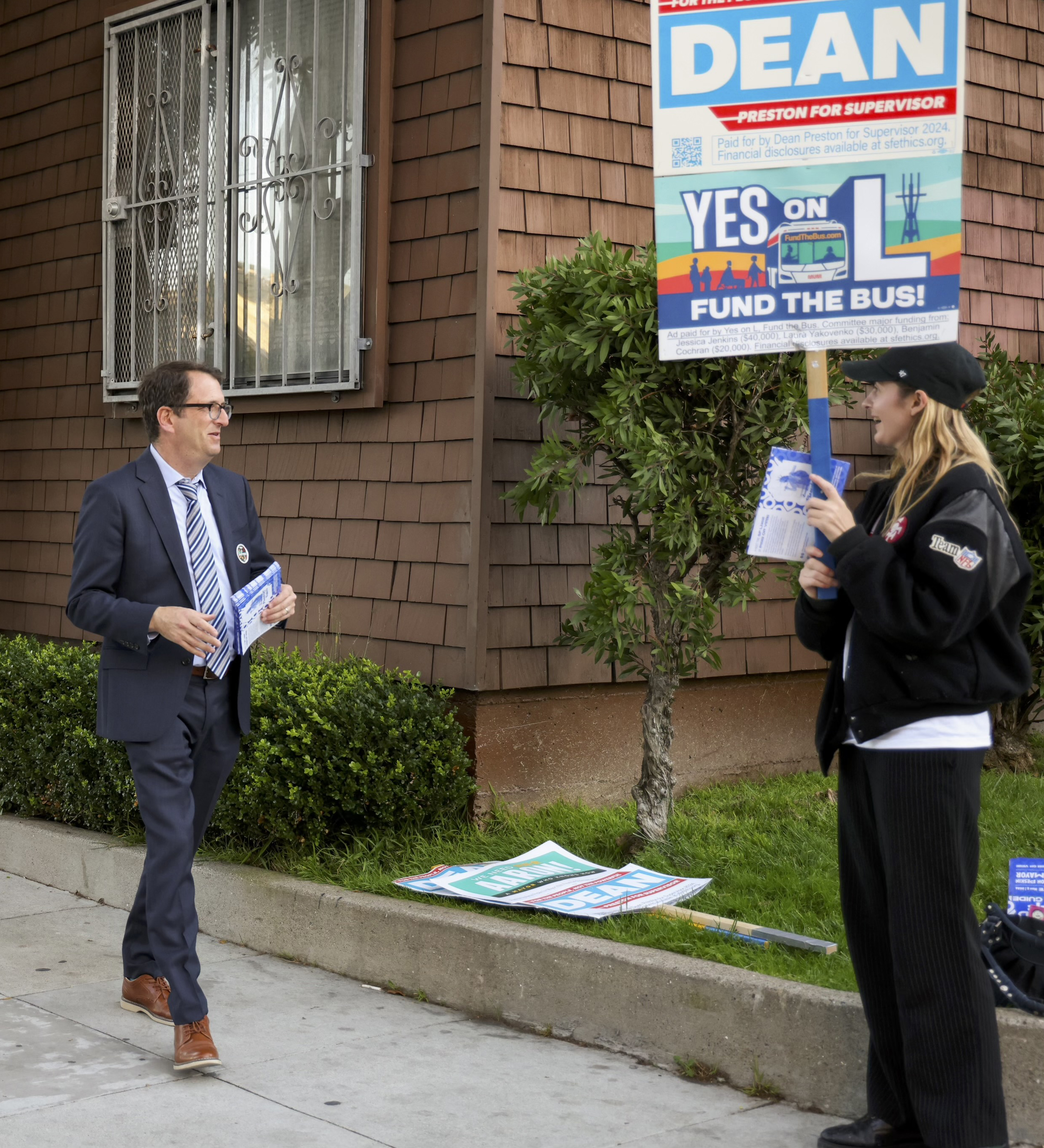 A person in a suit hands out flyers on a sidewalk, approaching another person with a large campaign sign advocating for funding public transportation.