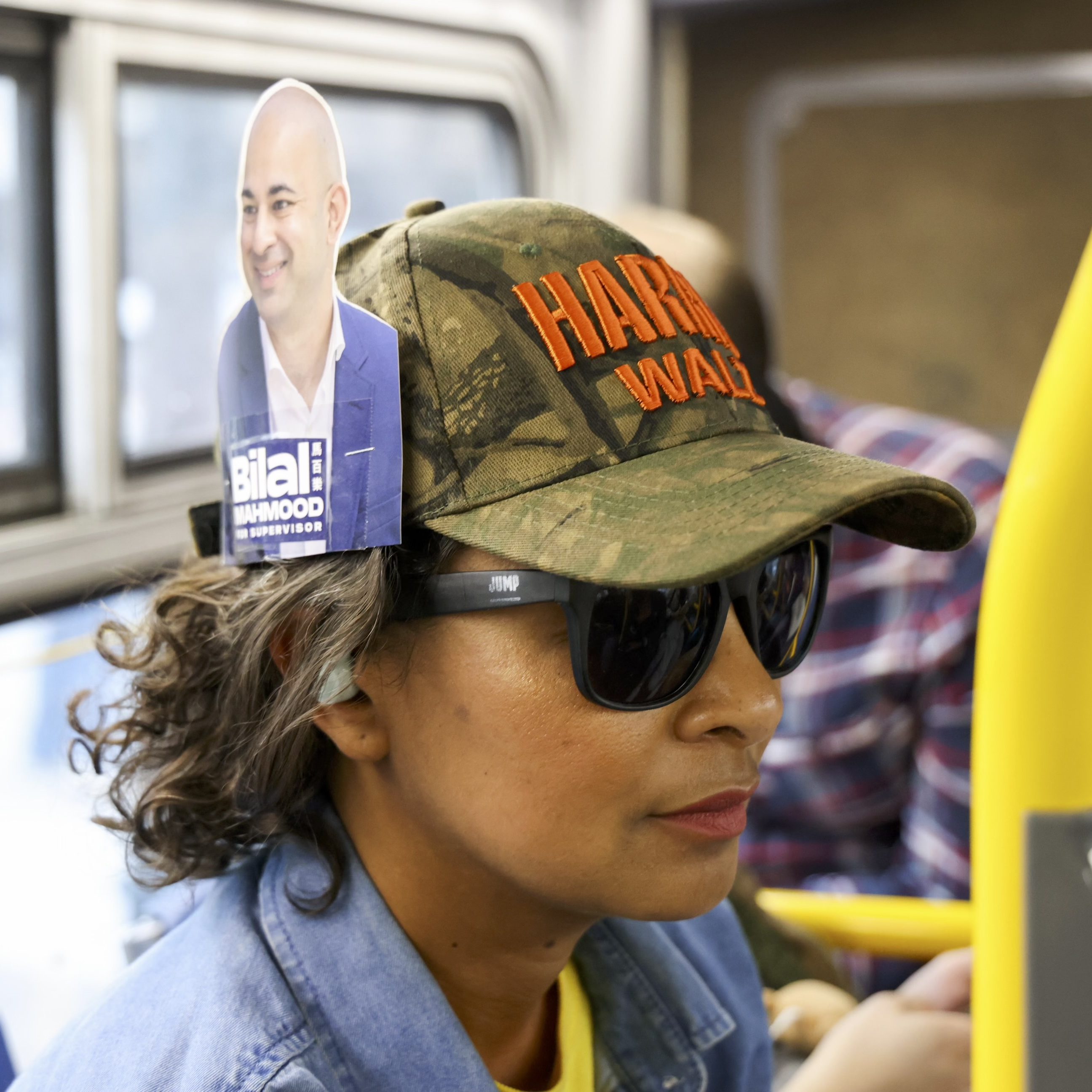 A person wearing sunglasses and a camo cap with an orange logo sits on a bus. A flyer with a photo of a smiling person is tucked into the side of the cap.