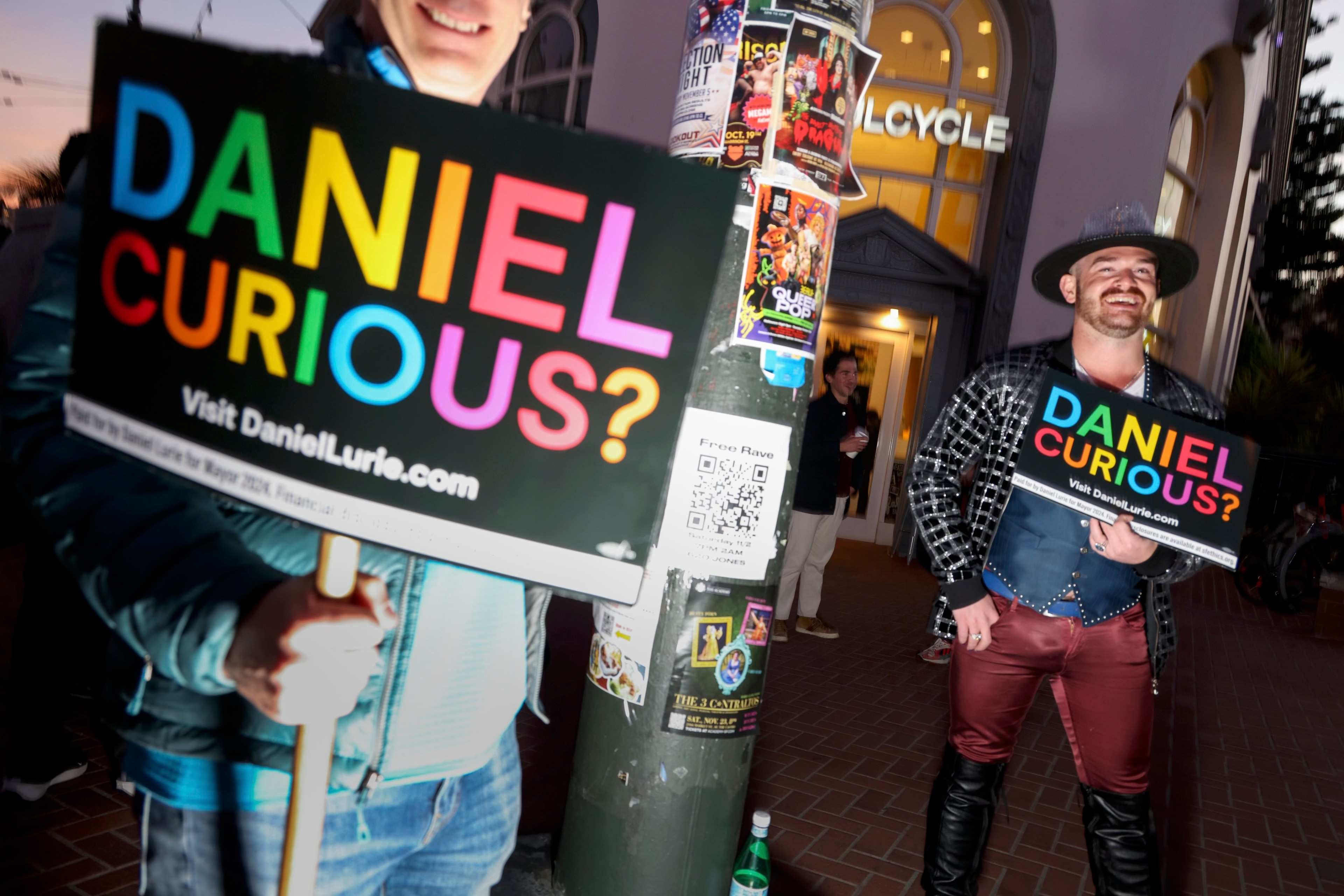 Two people hold colorful signs saying, &quot;DANIEL CURIOUS? Visit DanielLurie.com&quot; outside a venue. A pole is covered in event posters.