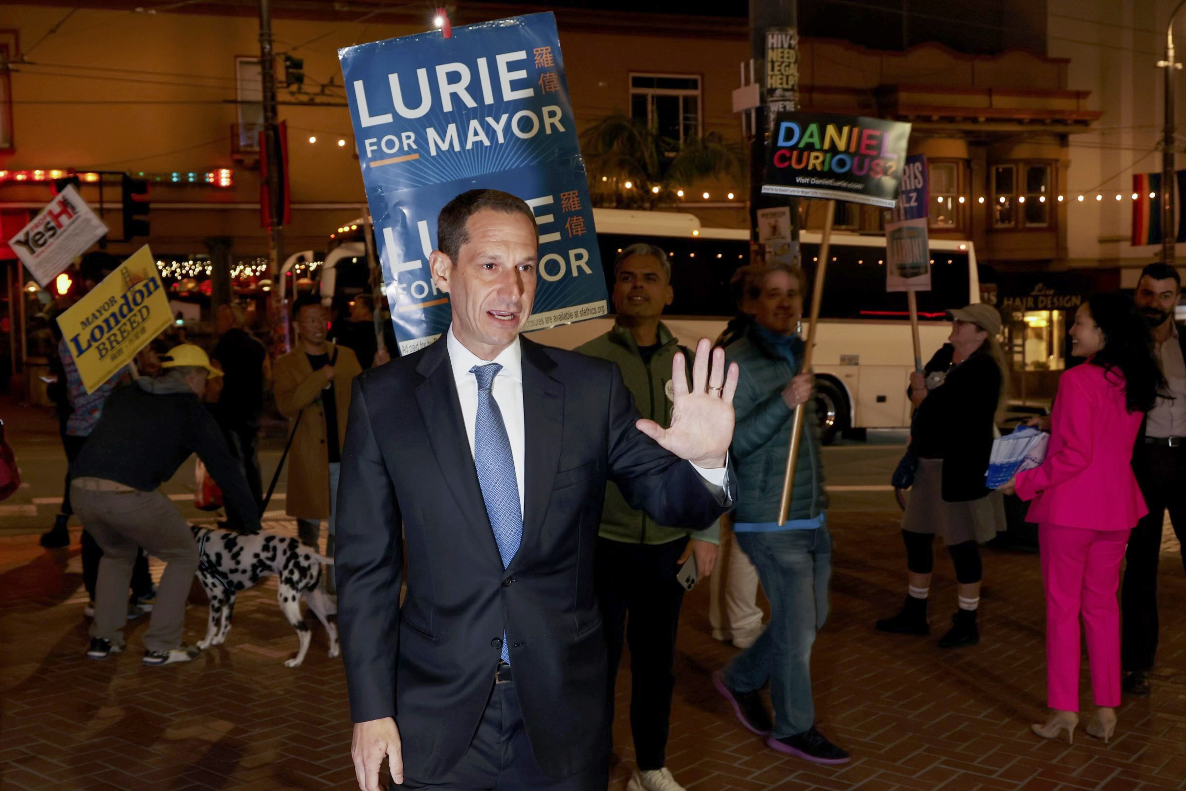 A man in a suit gestures to a crowd at night, surrounded by people holding political signs. There's a Dalmatian, and holiday lights are visible in the background.