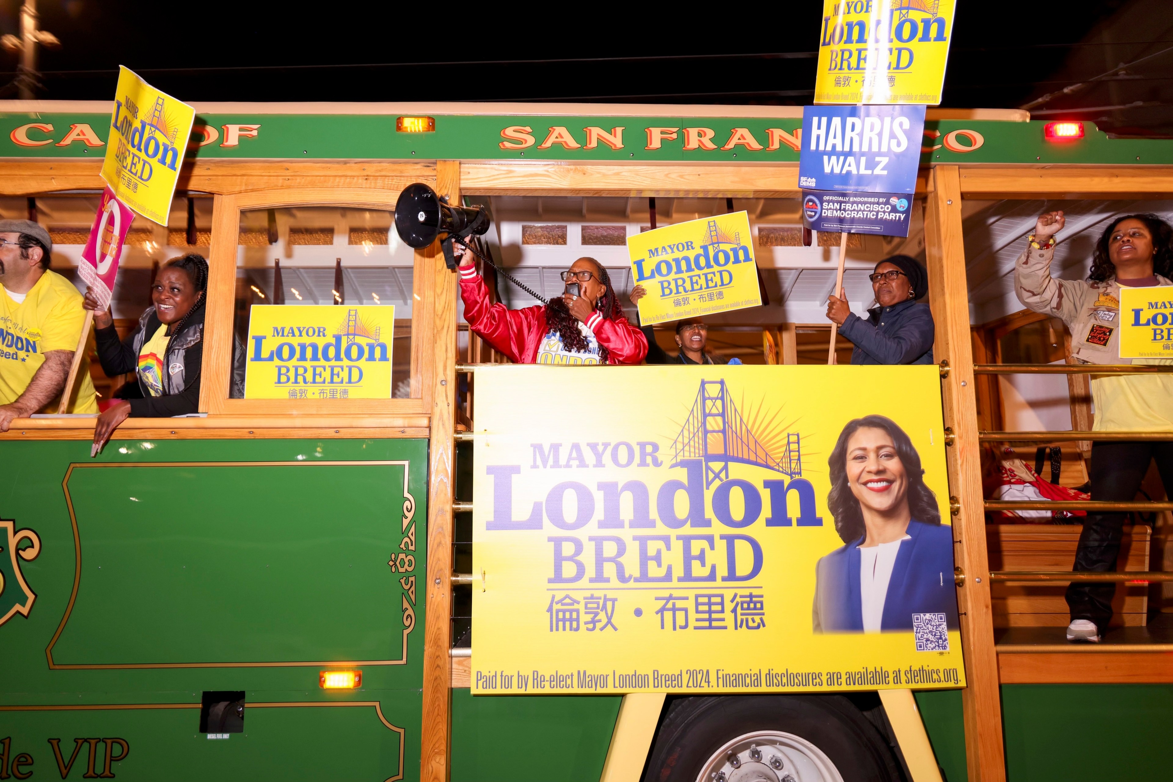 People on a San Francisco trolley hold campaign signs for Mayor London Breed, with a large banner displaying her image and name prominently.