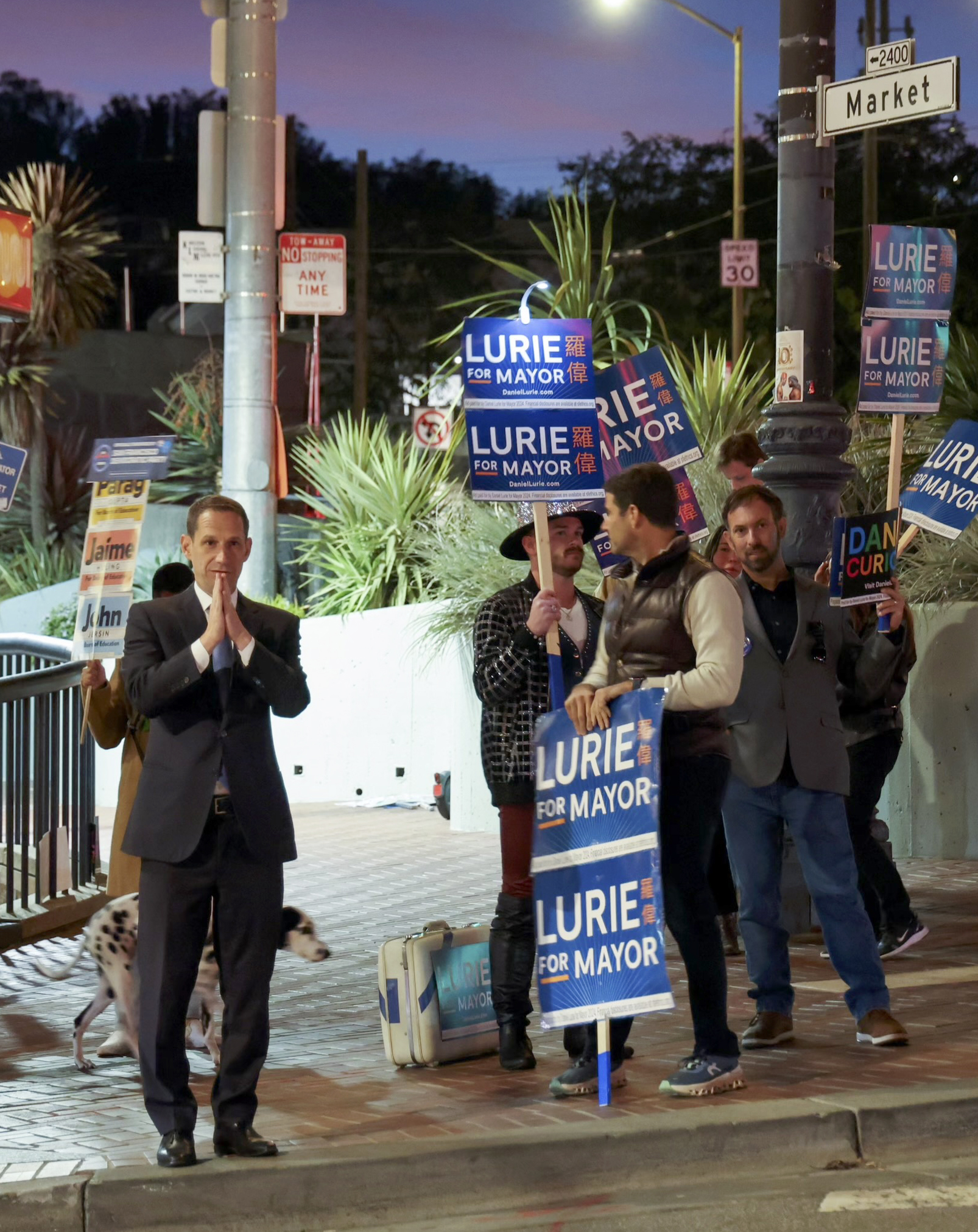 A campaign scene features a group of people holding &quot;Lurie for Mayor&quot; signs, standing on a street corner at dusk, with a man in a suit joining the gathering.