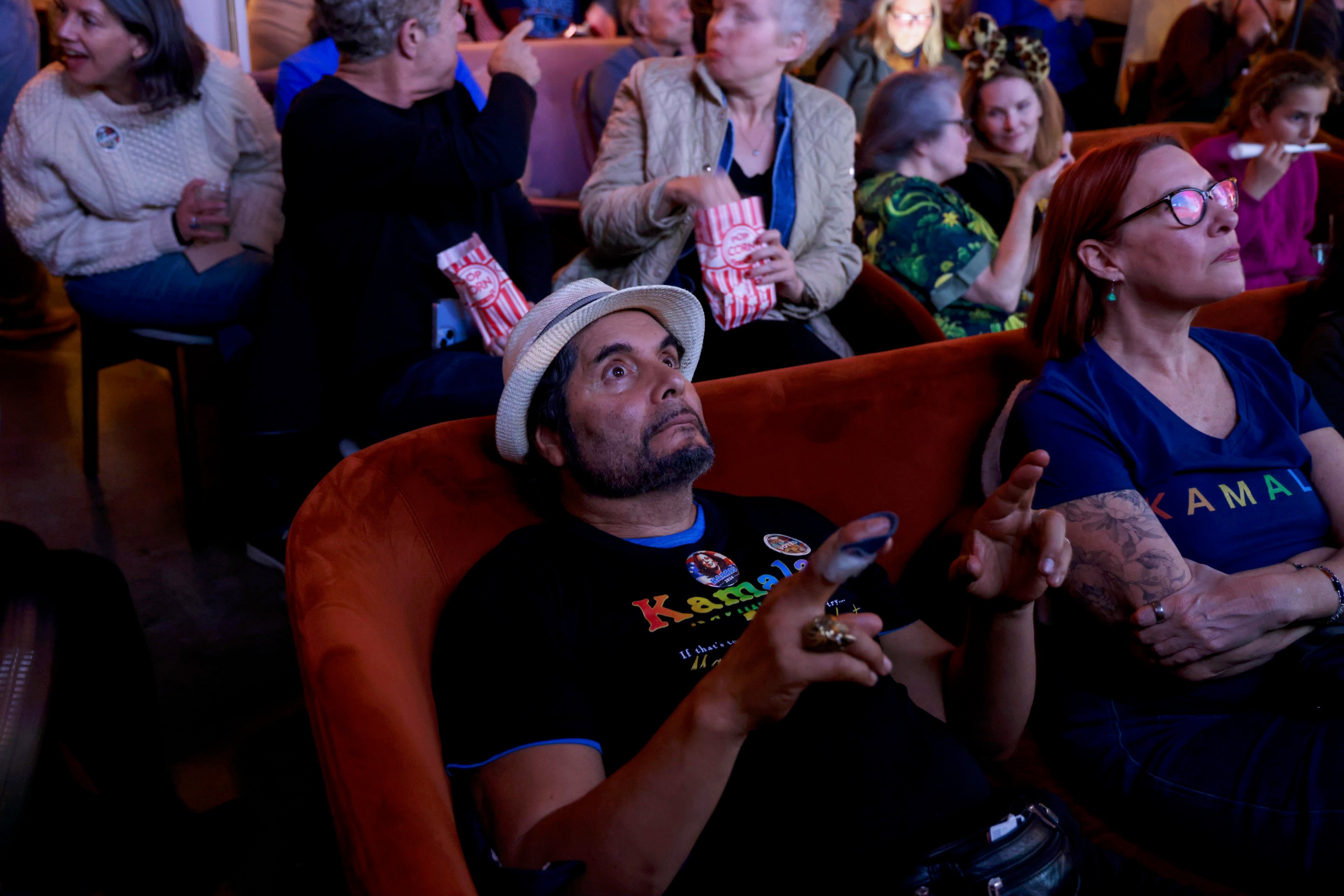 A man in a hat sits on an orange chair, holding a phone with his finger bandaged, while people around him chat and eat popcorn in a crowded, dimly lit room.