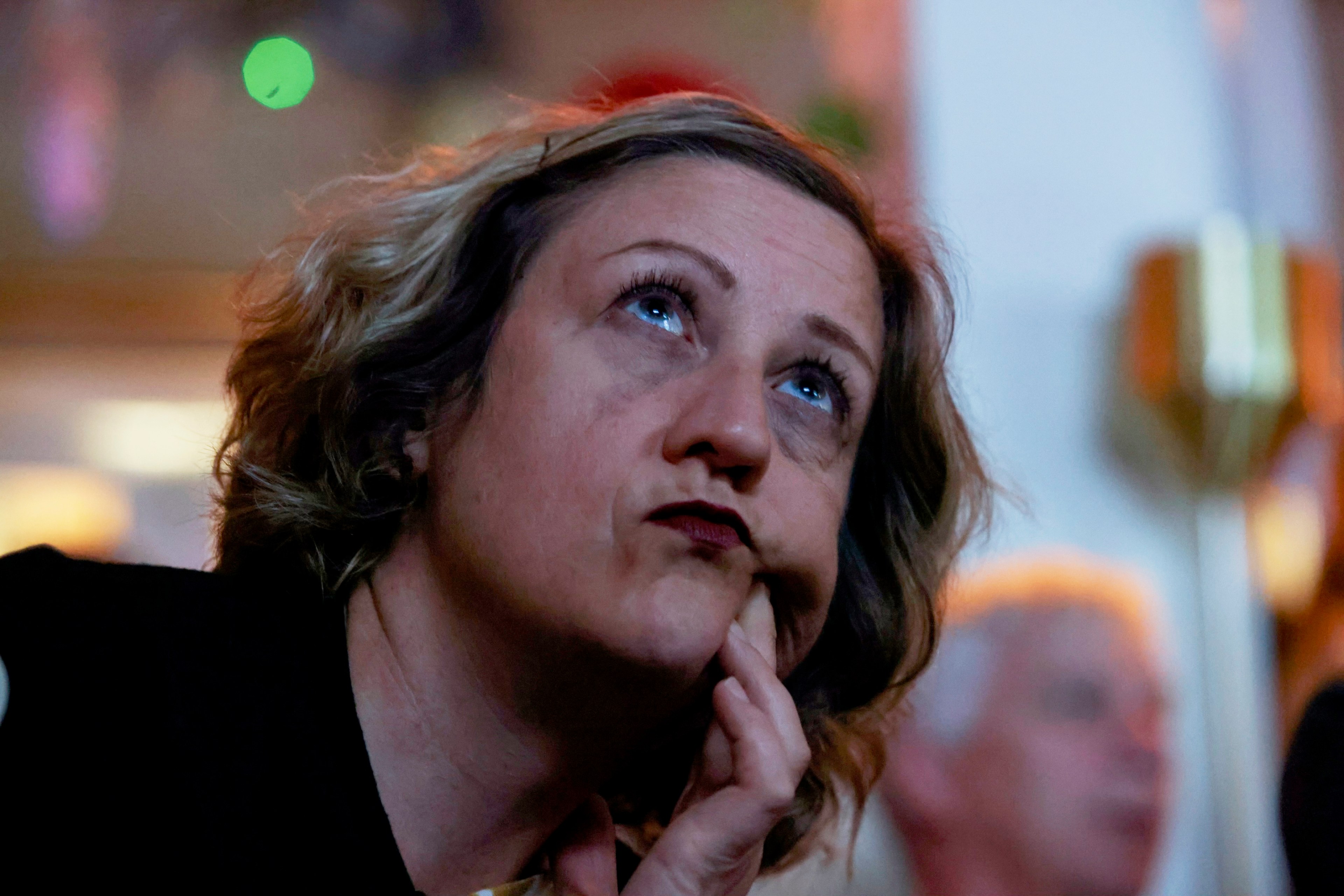 A woman with light hair gazes upward thoughtfully, her finger resting on her cheek. The background is softly blurred, with warm lighting and indistinct figures.