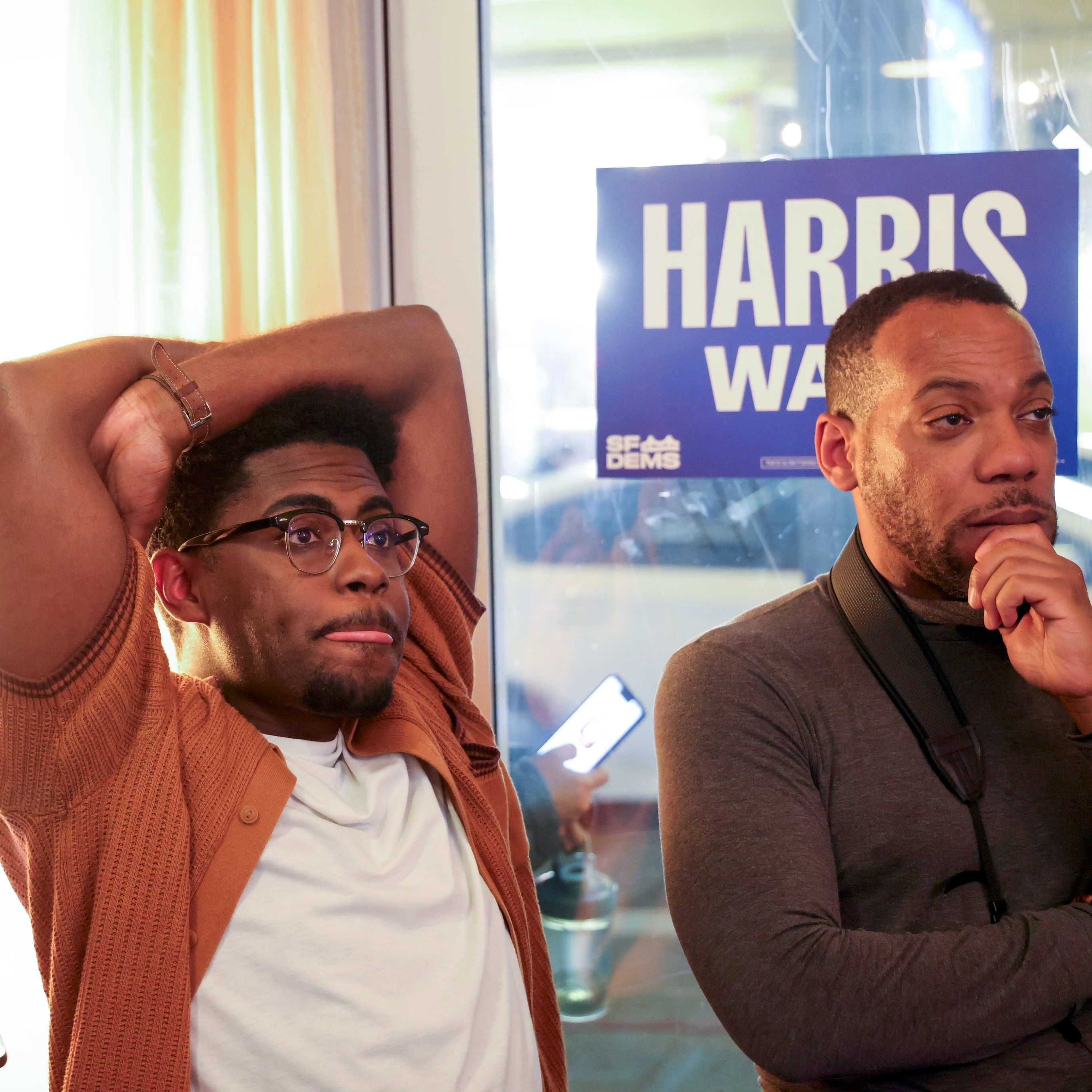 Three men are indoors, one checking his phone, another with hands on his head, and the third looking thoughtfully. A "Harris" sign is visible behind them.