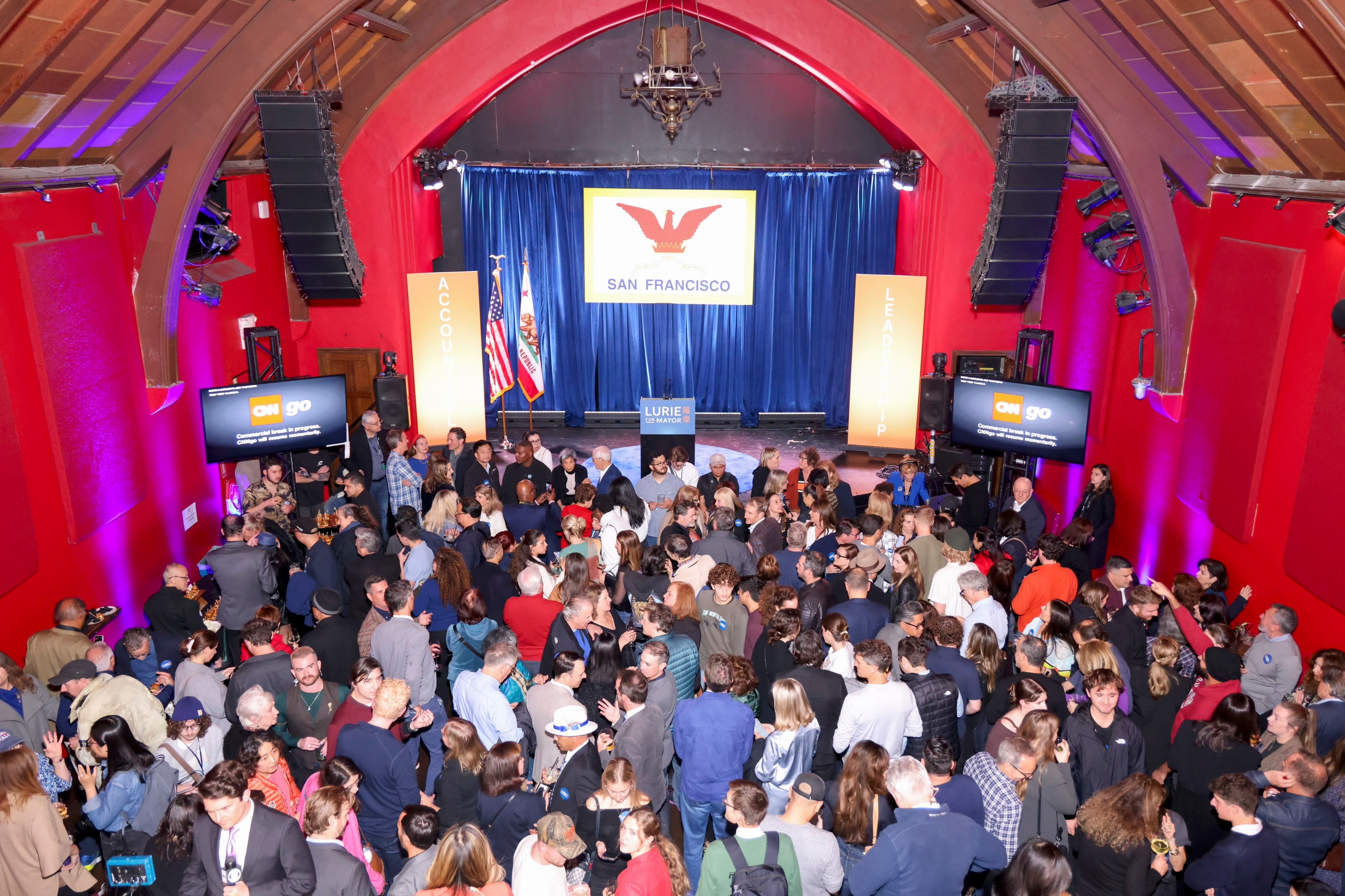 A large crowd gathers in an auditorium with red walls, watching a stage displaying &quot;San Francisco&quot; with two flags and a sign reading &quot;Lurie for Mayor.&quot;