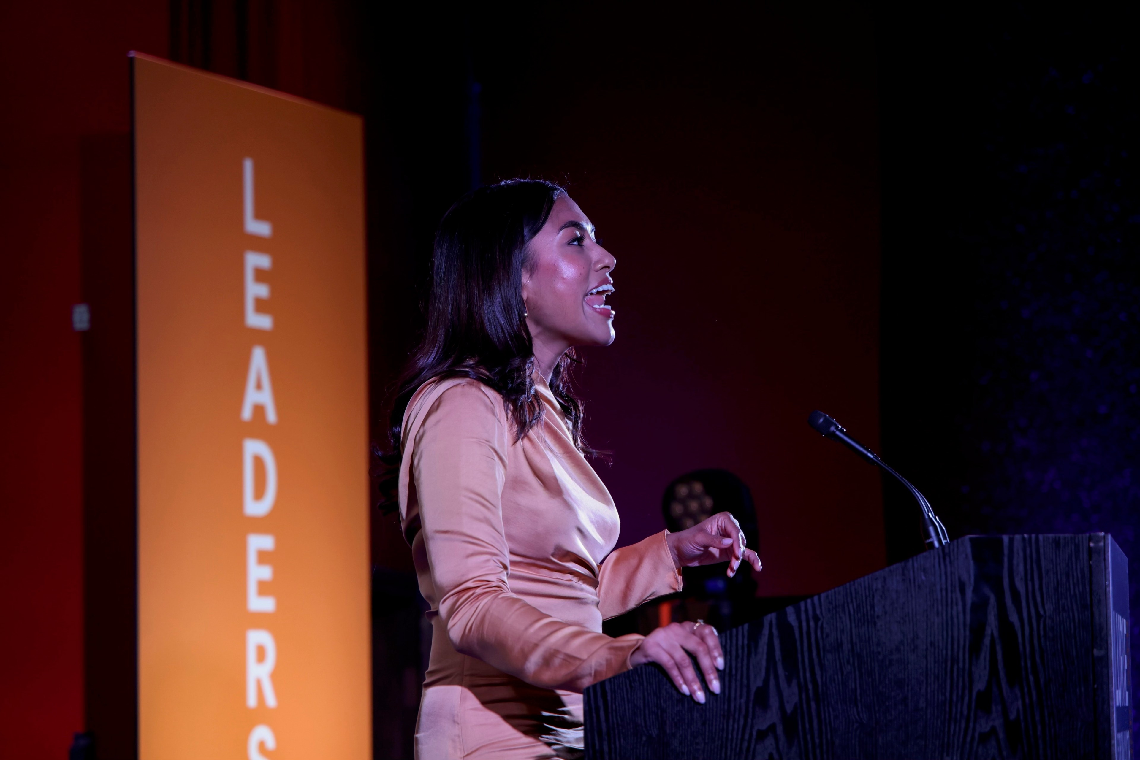 A woman in a gold dress is speaking at a podium with a microphone. Behind her is an orange sign with the word &quot;LEADERS&quot; in white text.