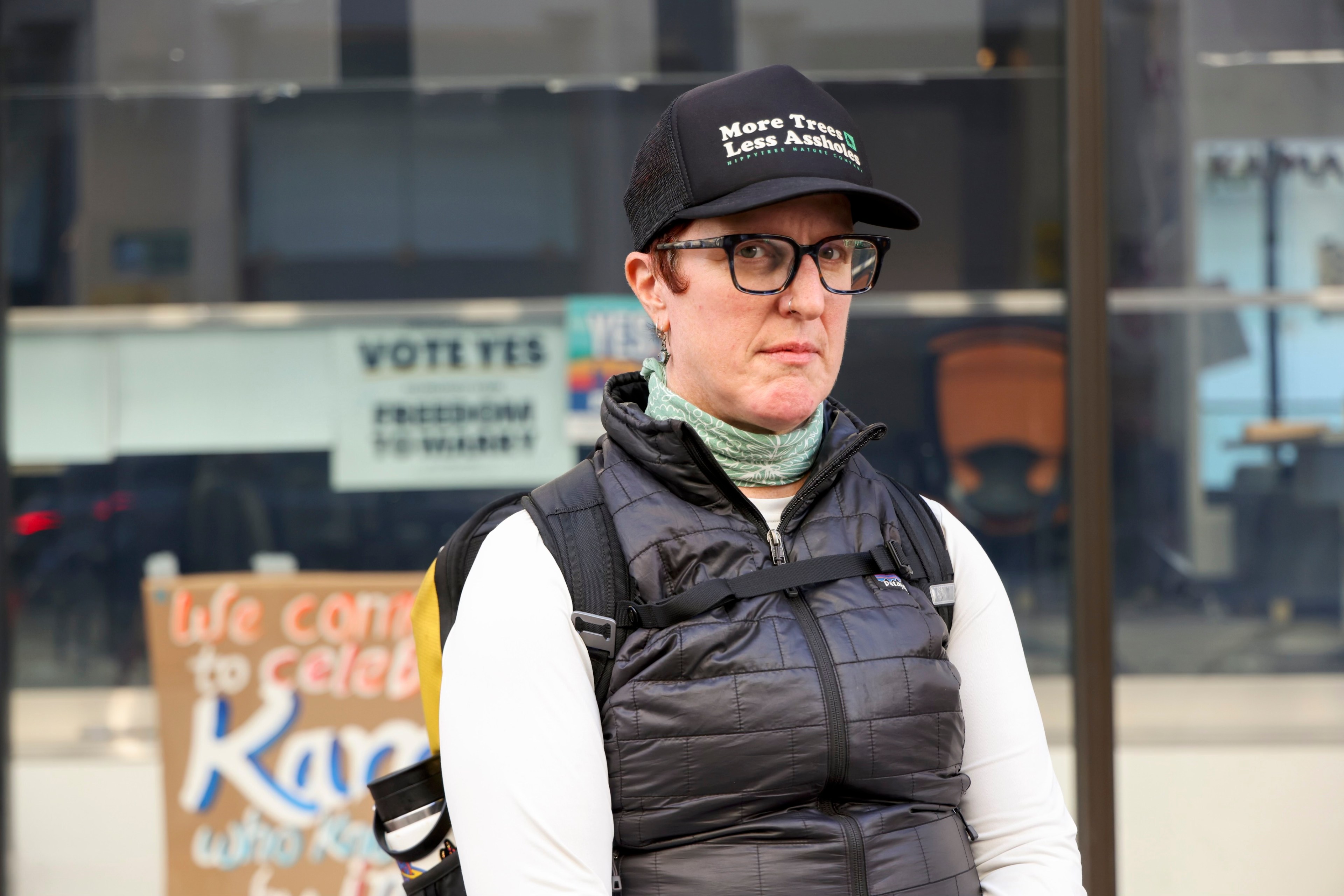 A person with glasses and a cap stands outdoors, wearing a black puffy vest and white shirt. Behind them is a sign reading &quot;VOTE YES&quot; and a partially obscured message on cardboard.