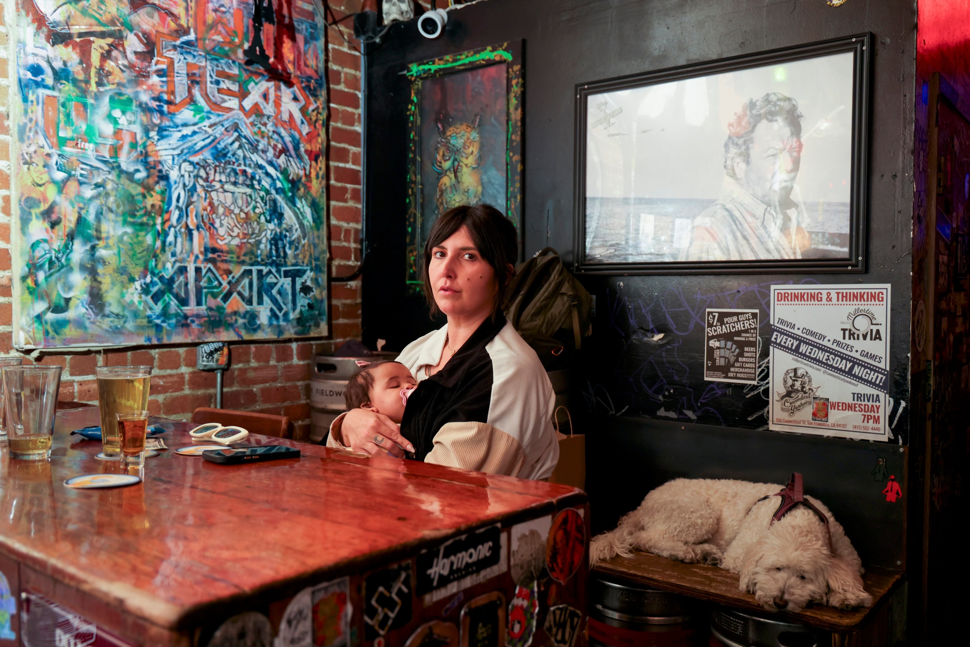 A woman holds a sleeping baby in a colorful bar with art on the brick walls. A fluffy dog sleeps on a bench beneath posters advertising trivia night.