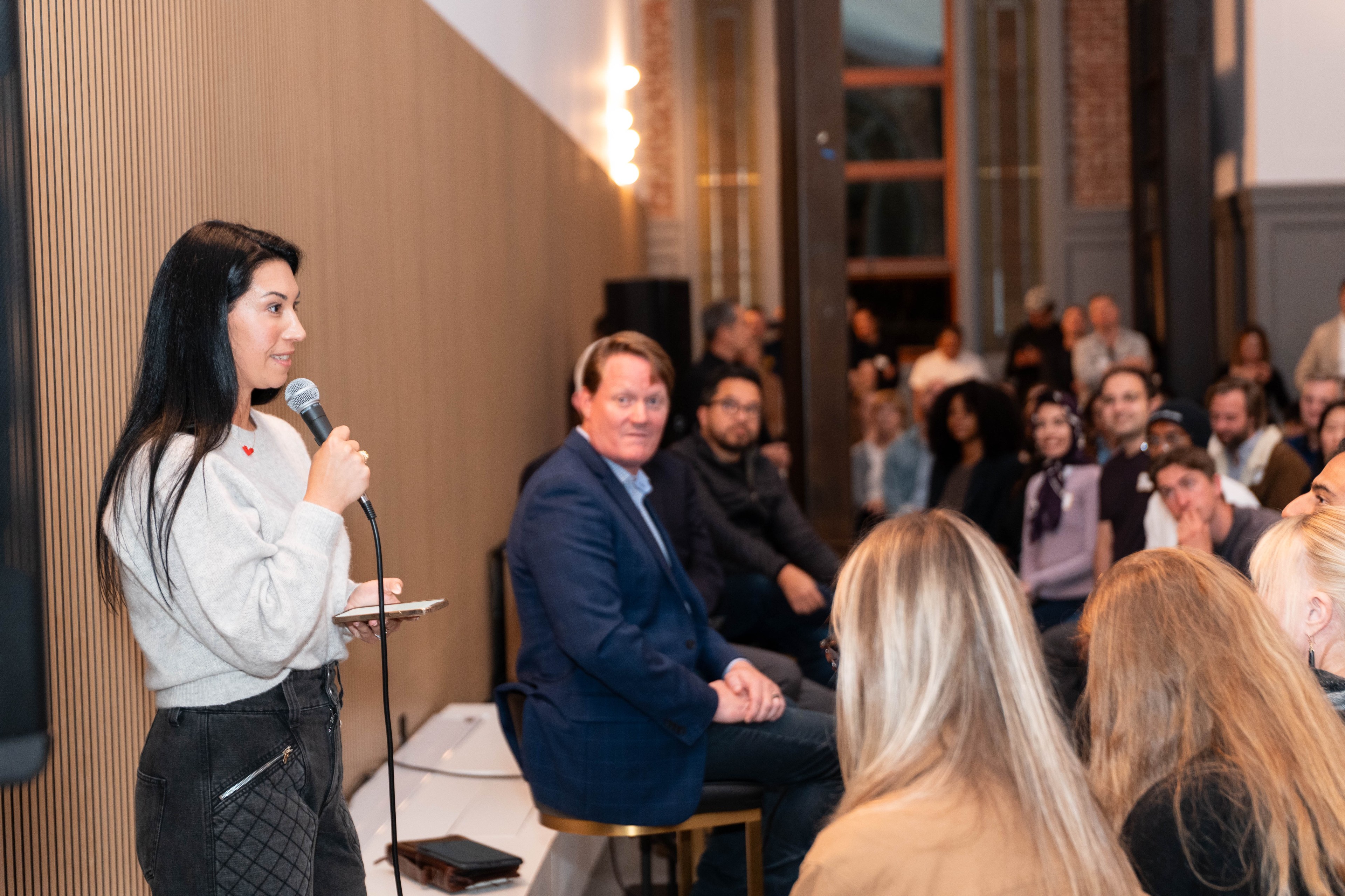 A woman holds a microphone, speaking to an audience in a room. People are seated, listening attentively with a visible man in a blue suit looking toward her.