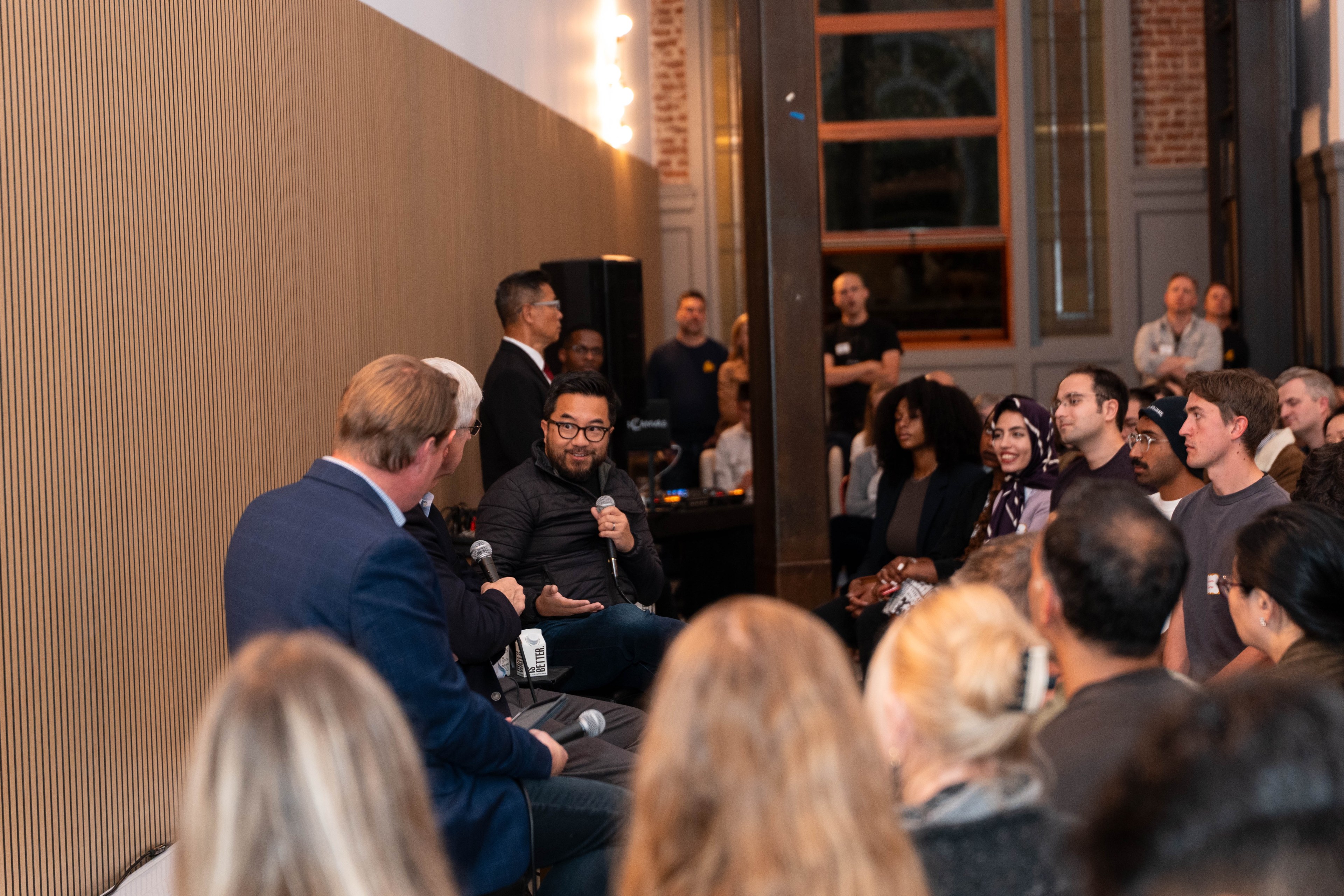 A group of people are sitting in a dimly lit room with wooden walls. One man with glasses is speaking into a microphone while others listen attentively.