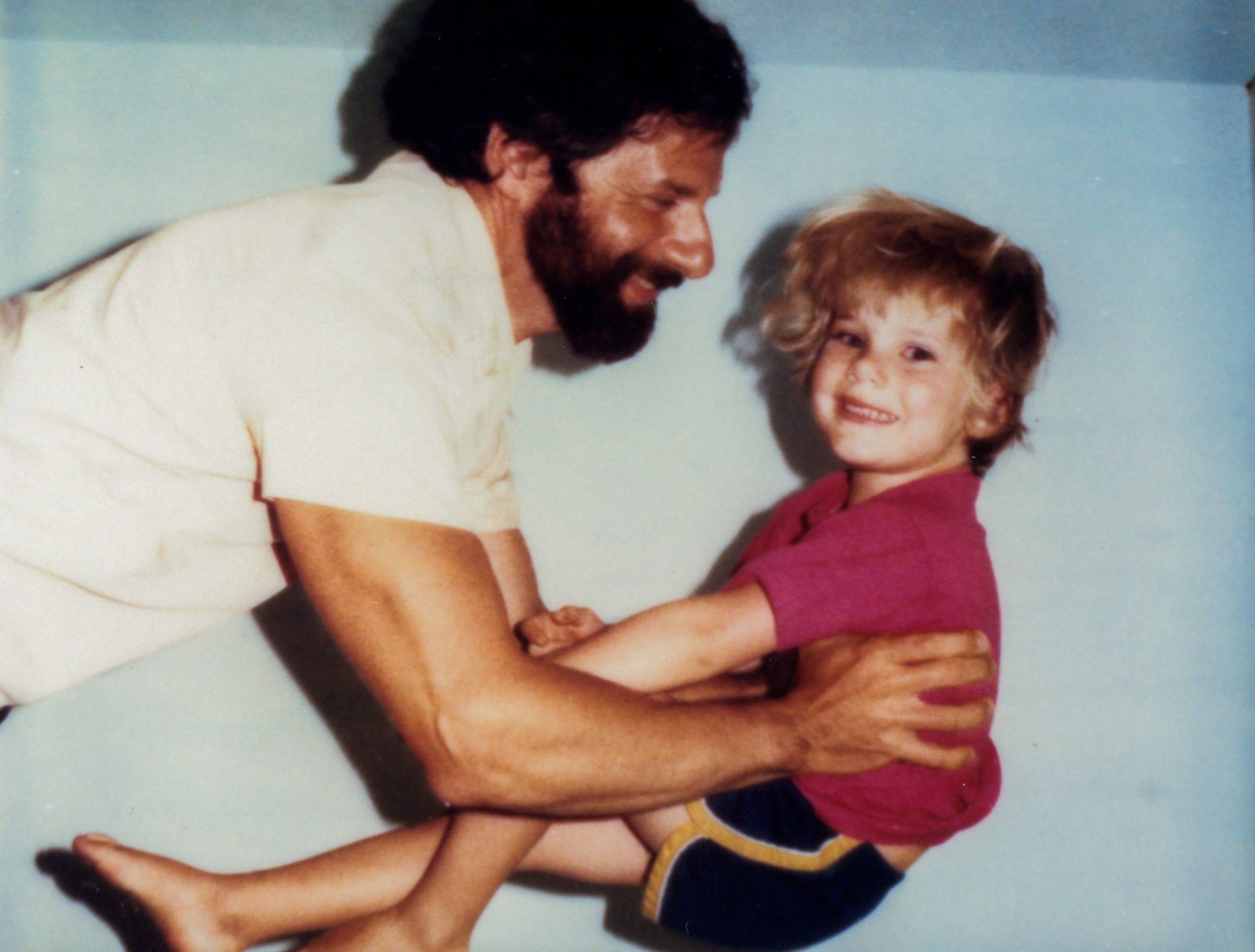 A man with a beard smiles at a young child he's holding playfully. The child, wearing a pink shirt and black shorts, is also smiling. The background is light blue.