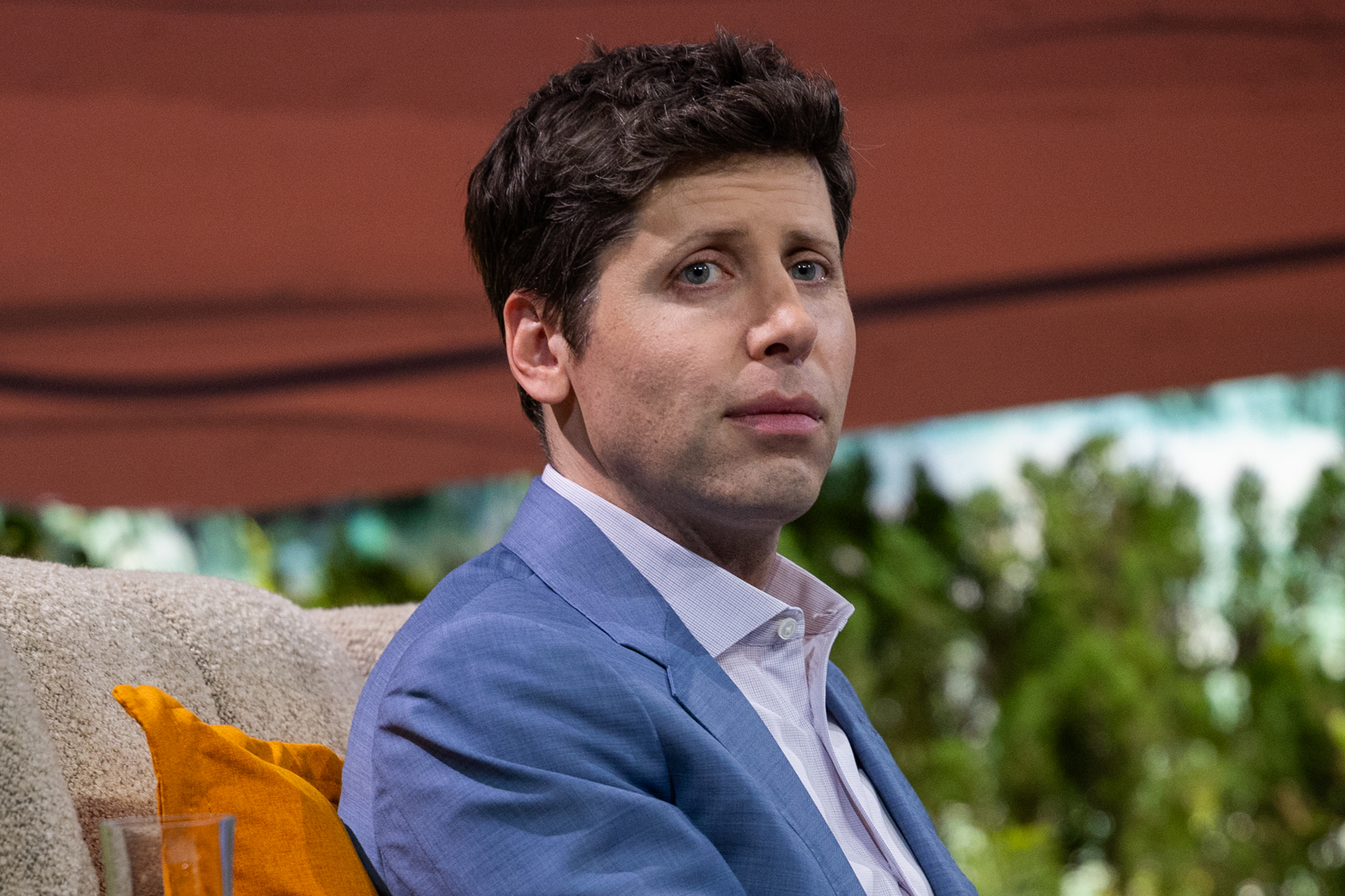 A person with short dark hair in a blue jacket sits on a cushioned seat. There is a yellow pillow behind them and a blurred green background.