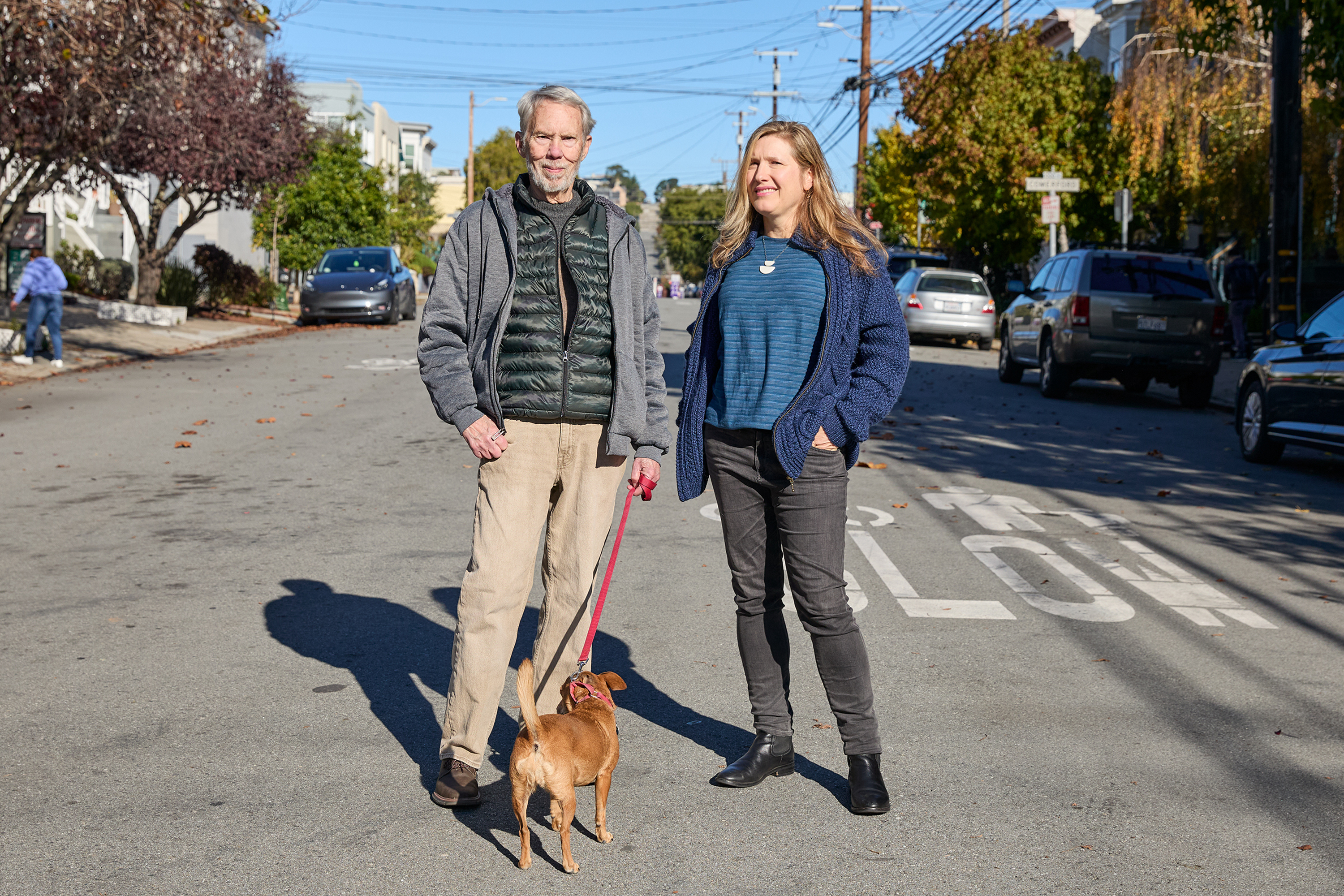 A man and woman stand on a sunlit street with a small dog on a leash. They are casually dressed, with trees and parked cars lining both sides of the street.