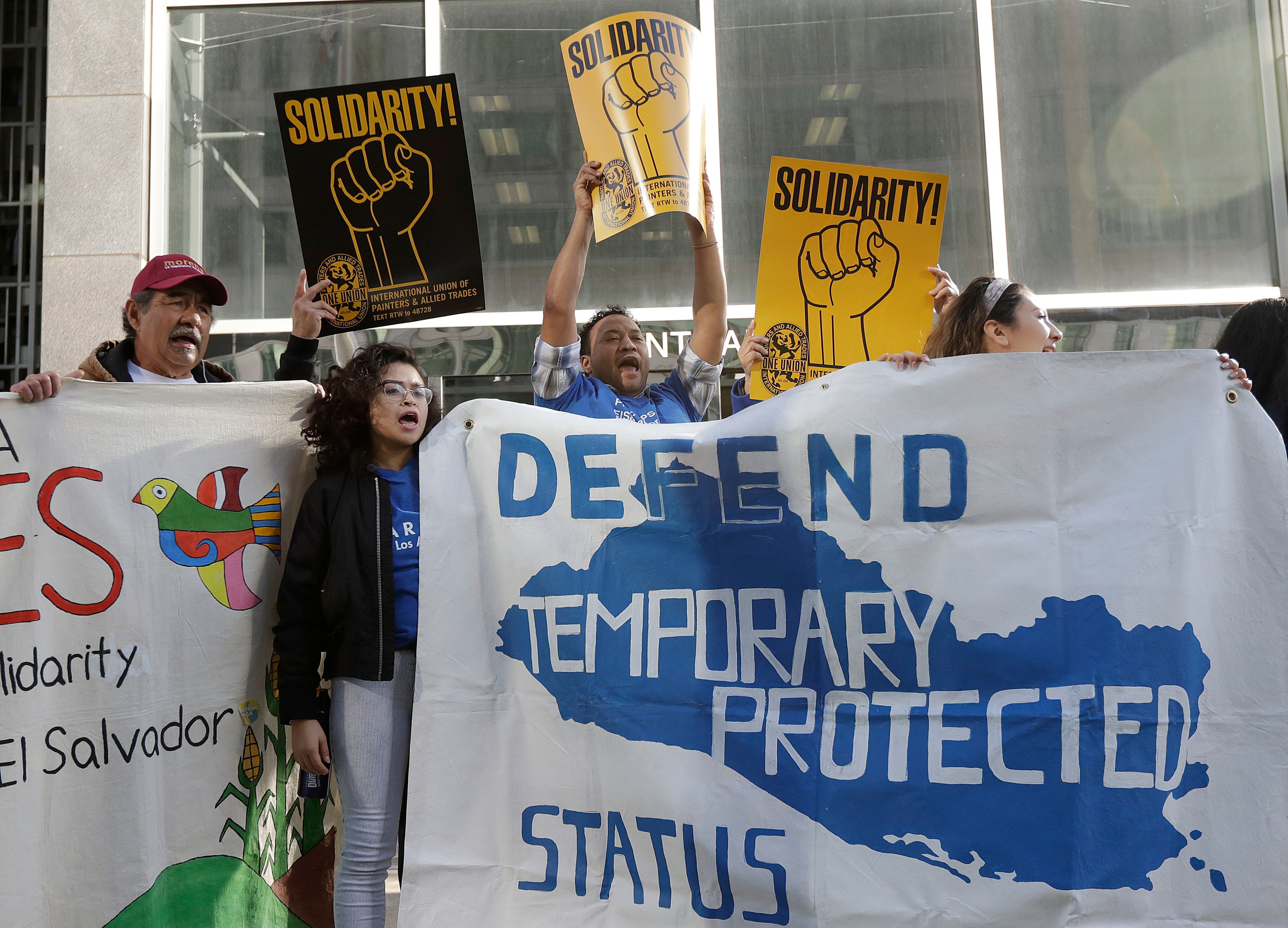 Protesters hold signs reading &quot;Solidarity!&quot; and a banner saying &quot;Defend Temporary Protected Status&quot; in bold text, amidst a backdrop of a building.